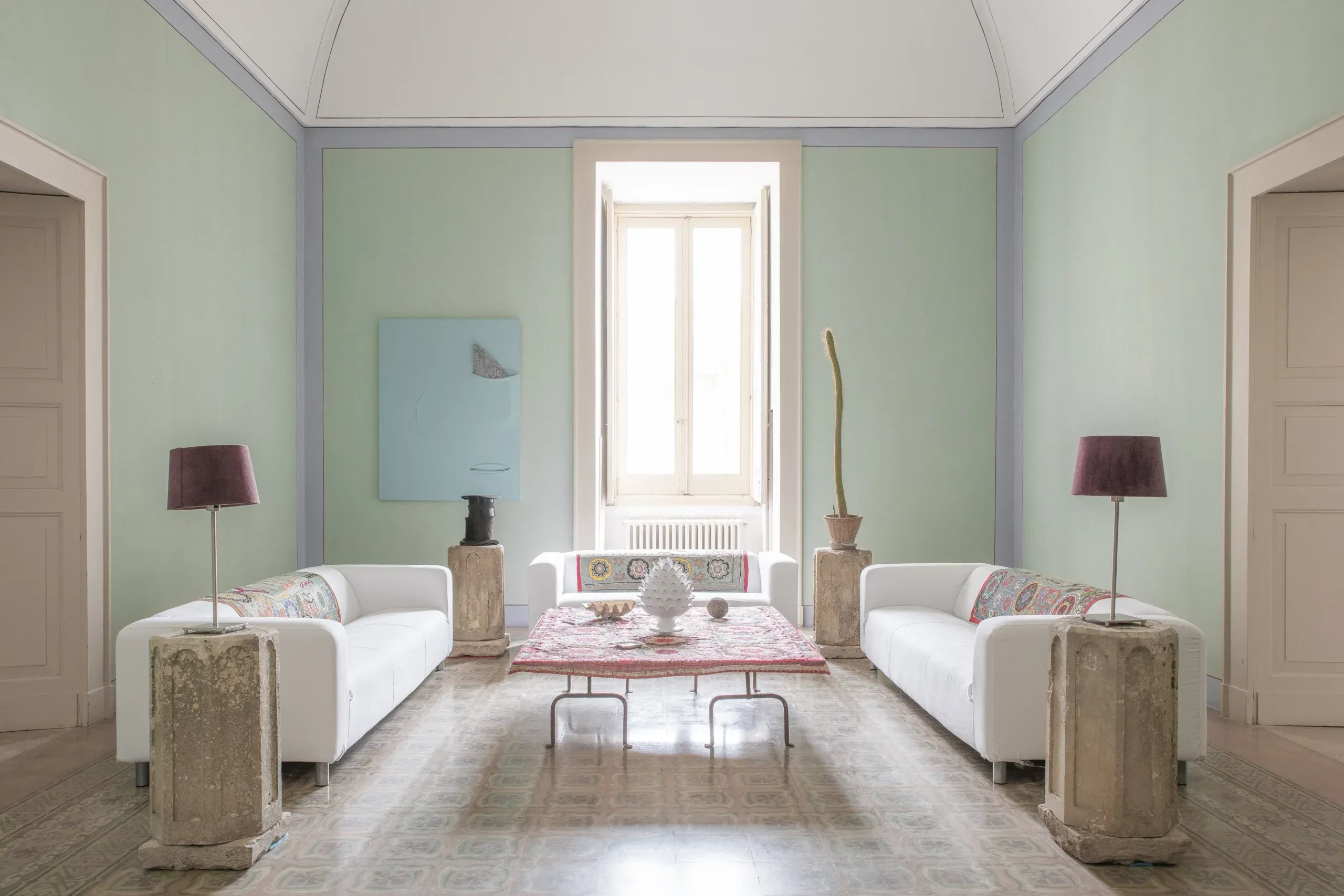 Palazzo Daniele, Europe — Symmetrical salon with pale green walls, white curved sofas, terracotta pedestals, burgundy lamps, and tall windows overlooking gardens.