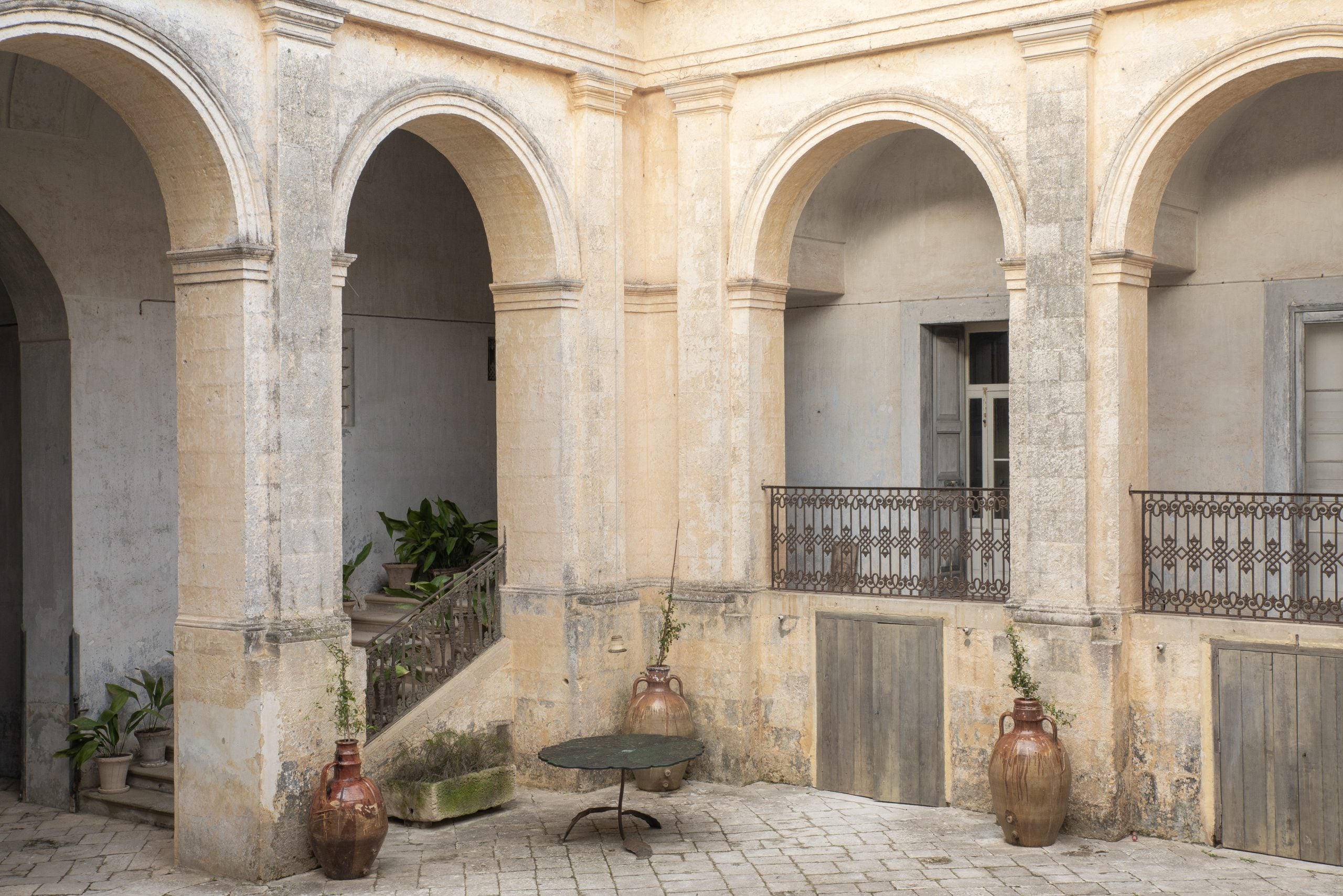 Palazzo Daniele, Europe — Neoclassical courtyard with arched colonnades, terracotta urns, worn limestone walls, ornate ironwork balconies, and dappled afternoon light.