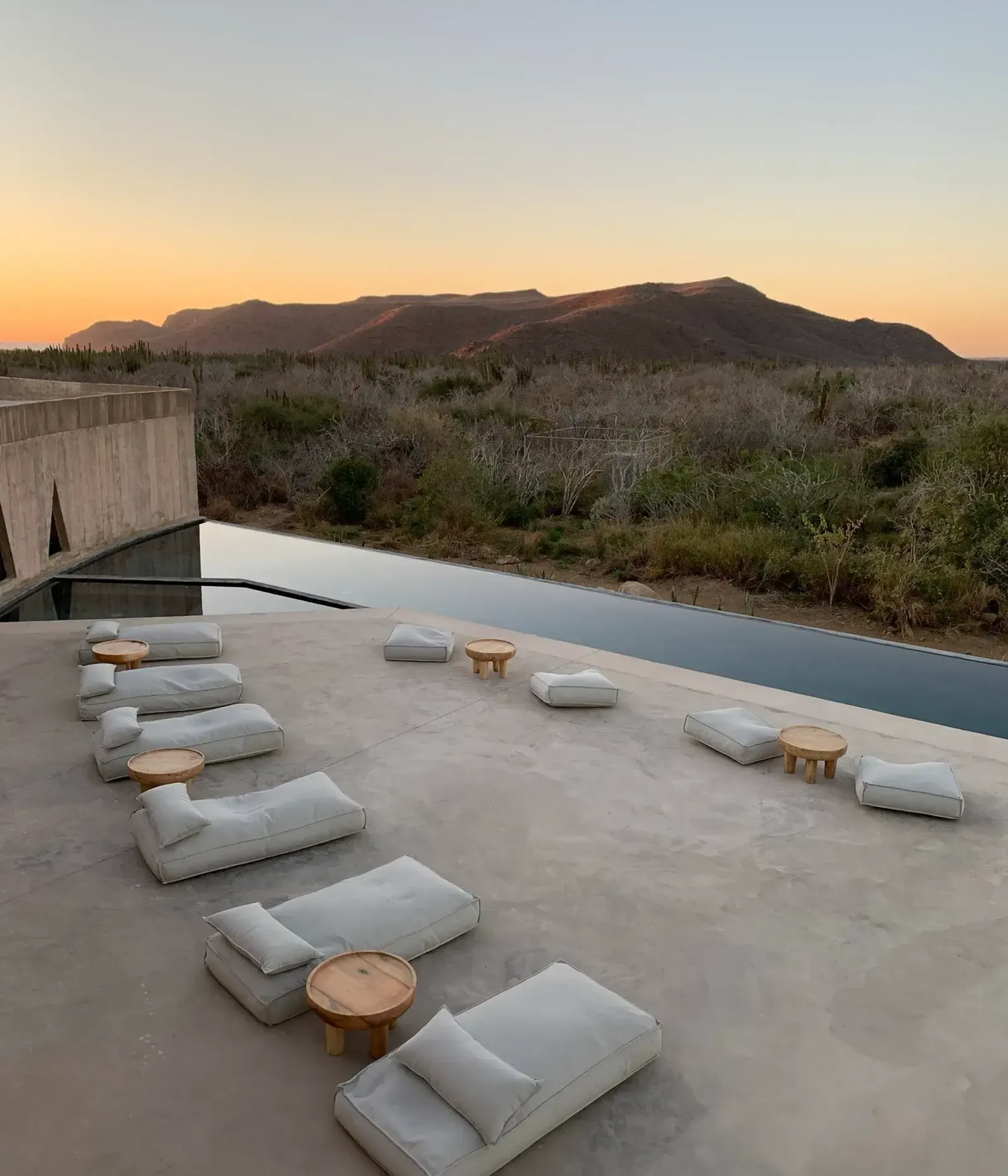 Paradero Todos Santos, Mexico — Concrete terrace with linen loungers and wooden side tables overlooking infinity pool, desert scrubland, and sunset-lit mountains at dusk.