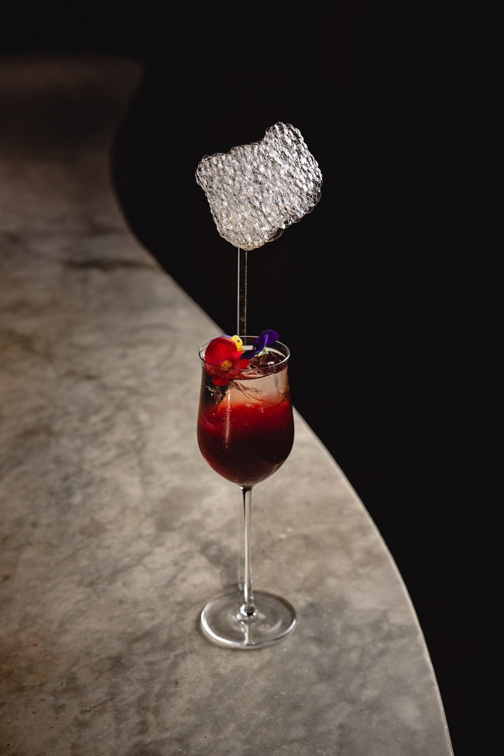 Paradiso Speakeasy, Europe — Ruby cocktail with ice and edible flower garnish, metallic leaf accent, dramatic shadow lighting on stone surface.