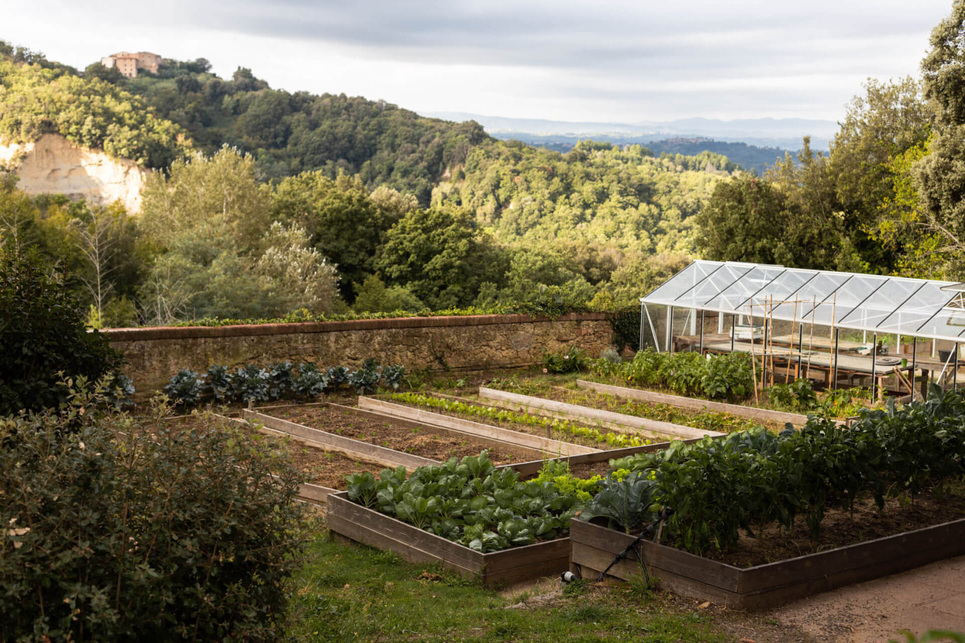 Villa Lena, Europe — Raised garden beds and greenhouse overlooking forested valley with stone fortification and distant hills.