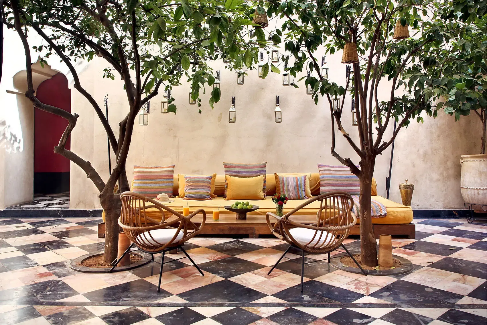 El Fenn, Marrakech, Morocco — Courtyard with cream-washed walls, mature trees, geometric tile floor, woven chairs, striped cushions, and hanging lanterns.