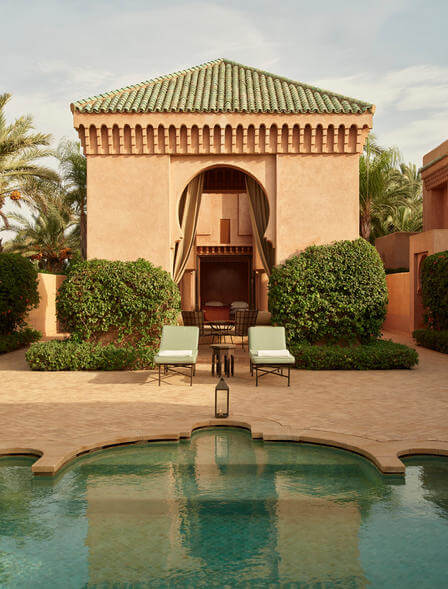 Amanjena, Marrakech, Morocco — Ochre-washed Moroccan pavilion with green-glazed tile roof, arched doorway, flanked by sculpted hedges, reflecting pool in foreground, palm trees, courtyard terracotta paving.