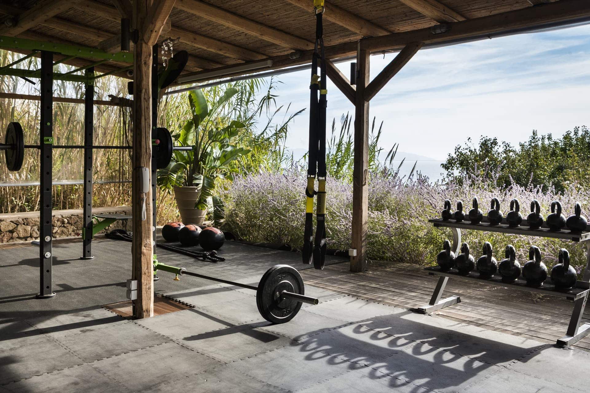 F Zeen Retreat, Europe — Outdoor fitness pavilion with wooden beams, barbell, kettlebells, and lavender garden views under open-air canopy.