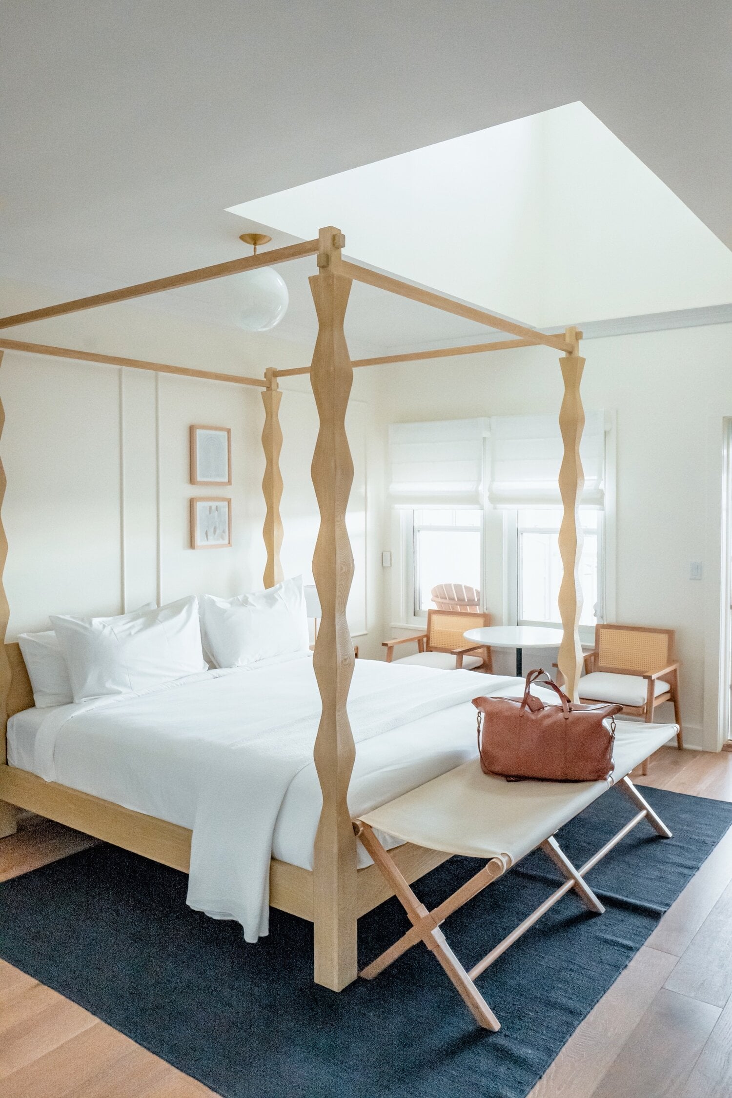 The Lake House, North America — Canopied bed with turned wooden posts, white linens, natural light from skylights, mid-century modern furnishings, minimalist luxury bedroom.