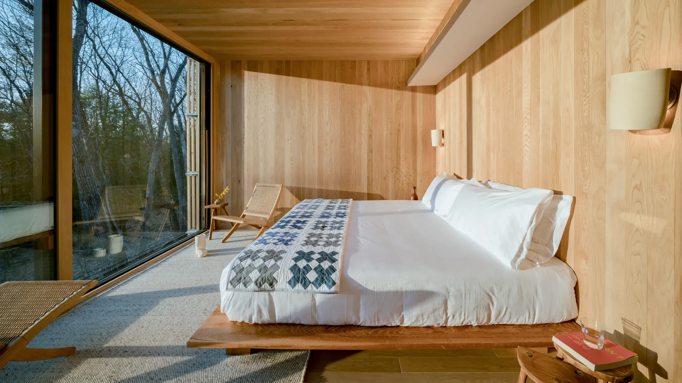 Piaule, North America — Minimalist wooden bedroom with platform bed, large windows overlooking bare trees, natural light, linen bedding, and mid-century furniture.