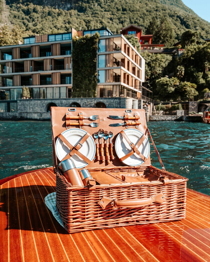 il Sereno Lago di Como, Italy — Open wicker picnic basket with leather straps on wooden deck overlooking turquoise Alpine lake and modernist waterfront architecture nestled in forested mountains.