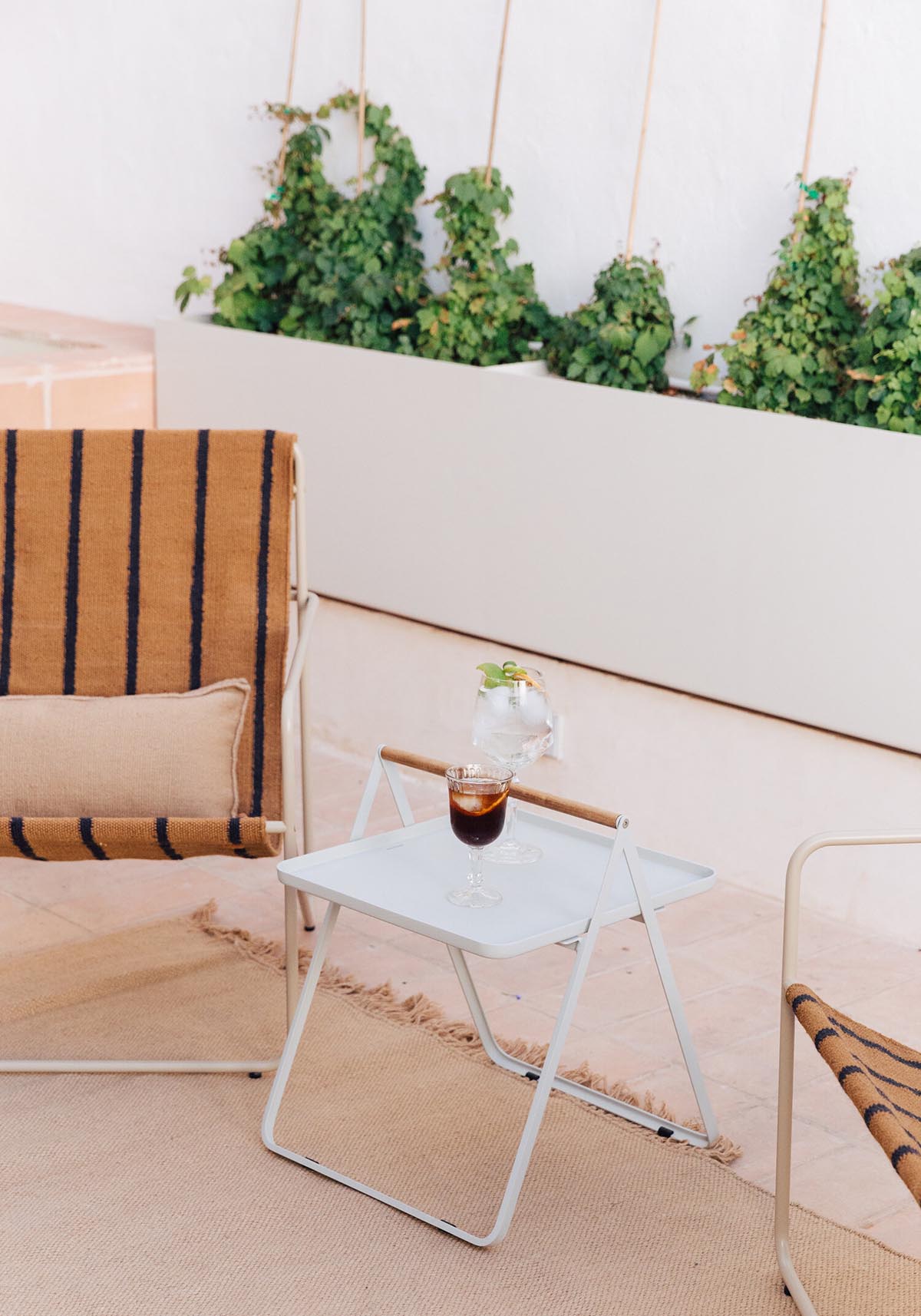 Plácido y Grata, Europe — Minimalist courtyard lounge with striped ochre chair, white side table, iced beverage, climbing ivy, and neutral palette.