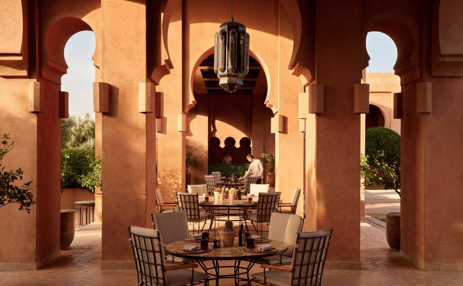 Pool Terrace & Olive Grove, Africa — Ochre arched colonnade with hanging lantern, terracotta courtyard, and dining tables overlooking gardens in warm sunlight.