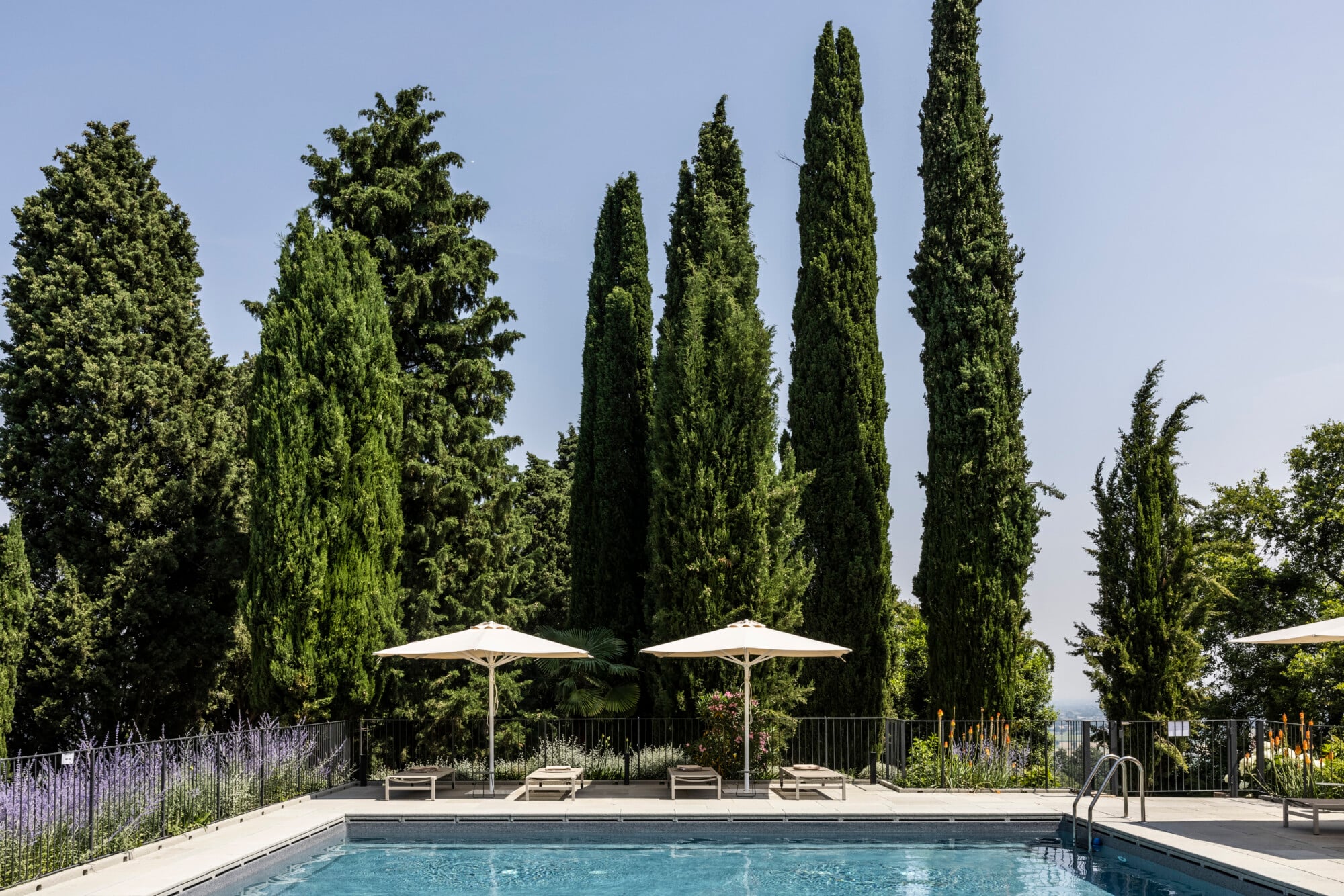 Roncolo 1888, Europe — Resort pool with loungers and umbrellas framed by tall cypress trees and manicured gardens under clear sky.