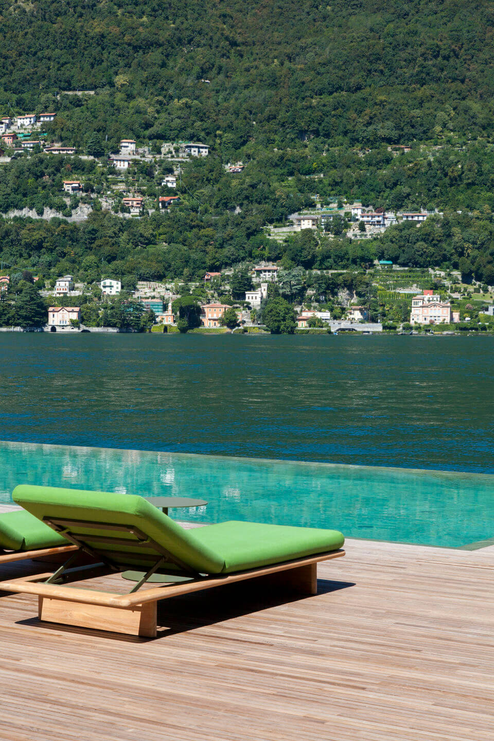 il Sereno Lago di Como, Europe — Green lounge chairs on wooden deck overlooking turquoise pool and forested lakeside village across deep blue water.