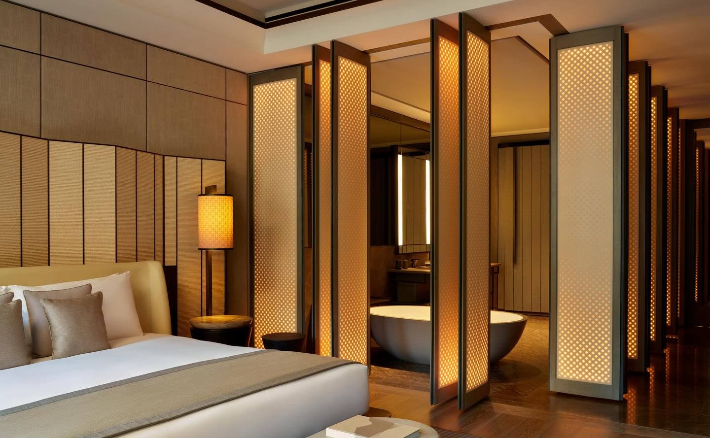 Aman New York, Manhattan — Luxury hotel bedroom with illuminated wooden lattice screens, contemporary furnishings, and spa-inspired ensuite bathroom visible beyond partition.