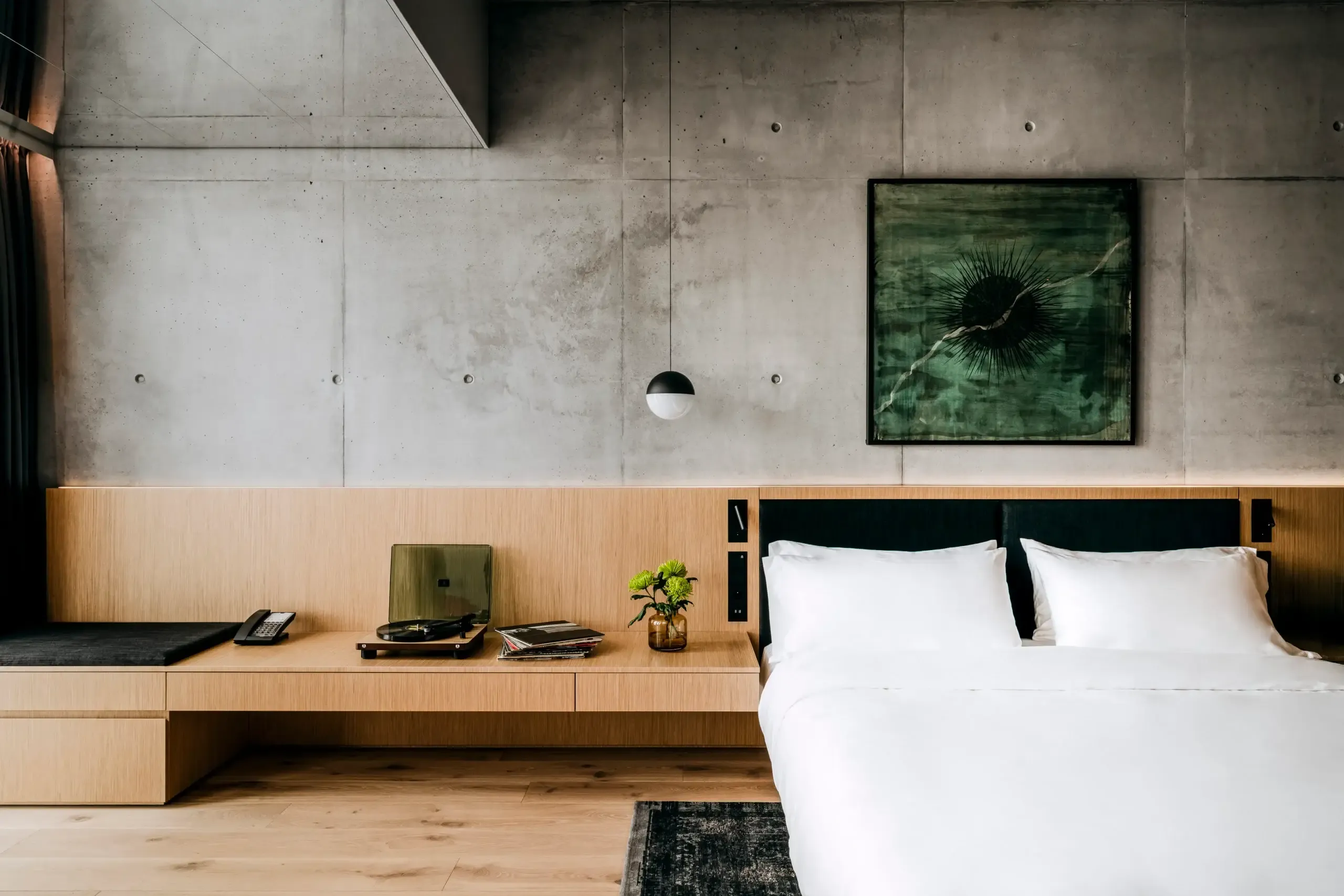 Nobu Warsaw, Poland — Minimalist hotel bedroom with light wood built-in desk, concrete walls, white bedding, and abstract green artwork above bed.