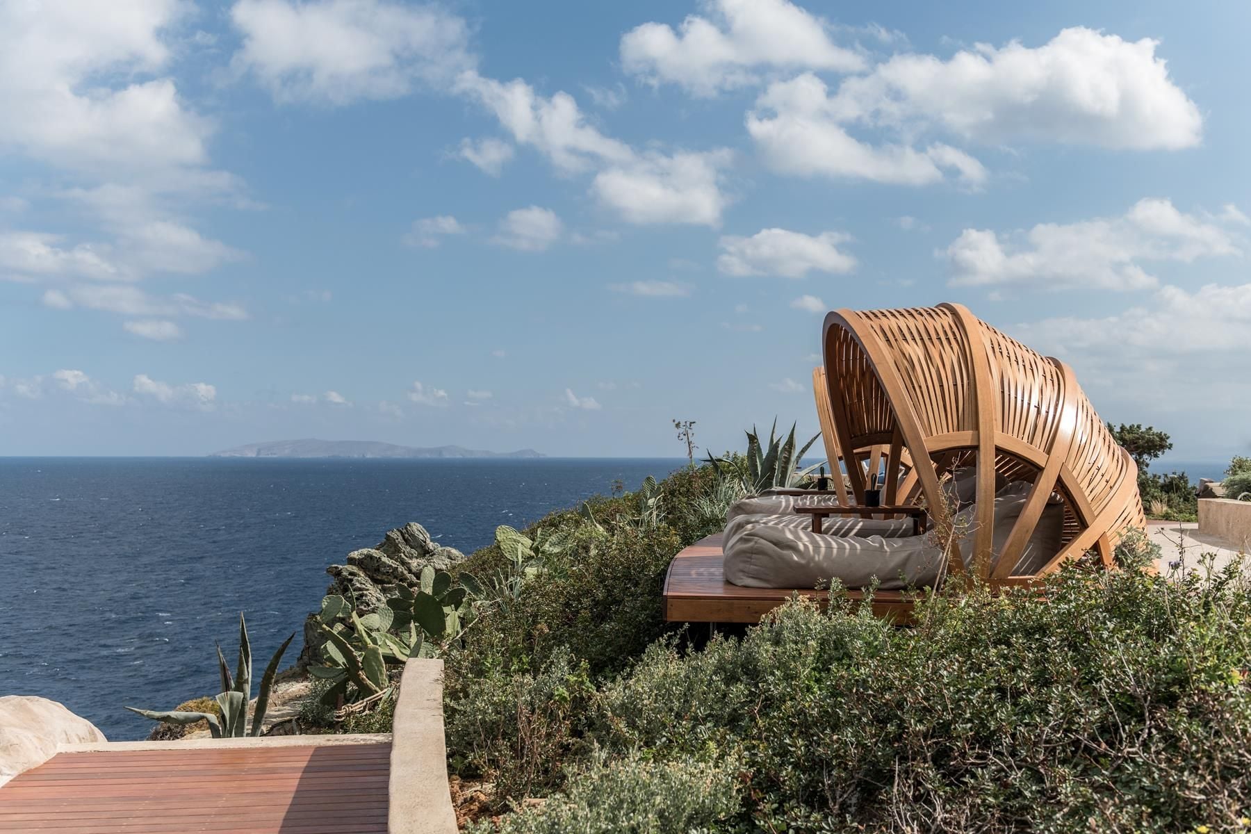 ACRO Suites, Europe — Wooden canopy bed with draped fabric overlooks Mediterranean coastline, surrounded by Mediterranean vegetation and azure sea under bright sky.