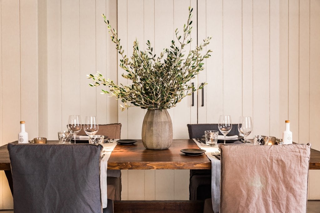 Sant Francesc Hotel Singular, Balearic Islands — Minimalist dining setup with wooden table, sage green floral arrangement, linen chair covers, and neutral aesthetic against paneled wall.