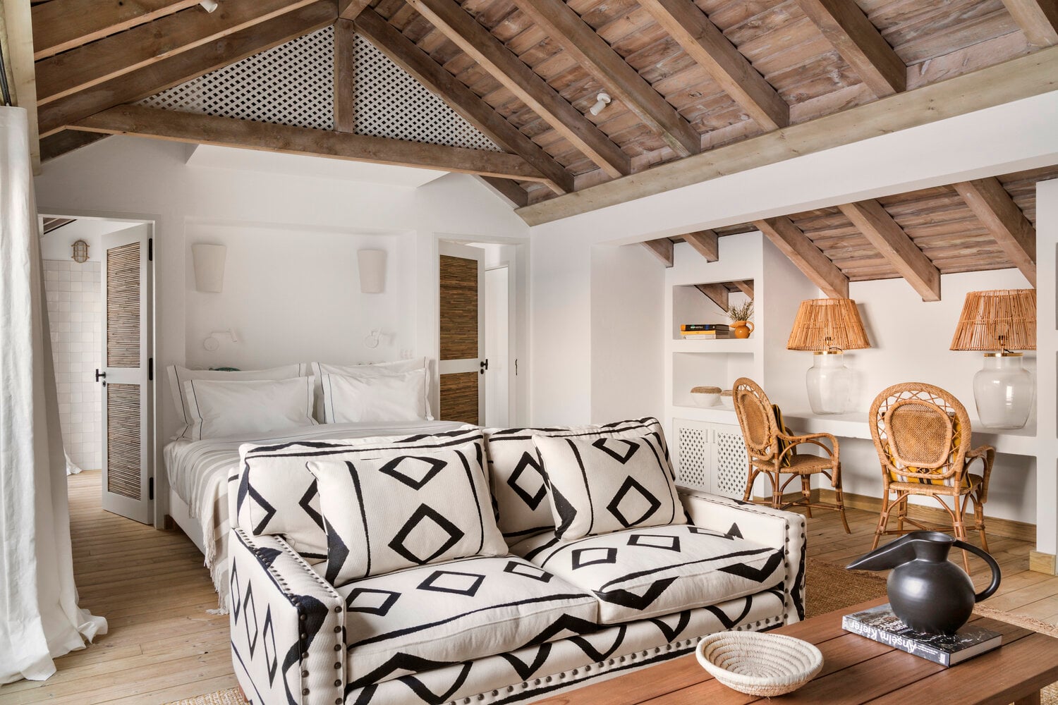 Quinta da Comporta, Europe — Rustic timber ceiling beams above modern bedroom with geometric patterned sofa, rattan chairs, and white walls in luxury Portuguese resort interior.