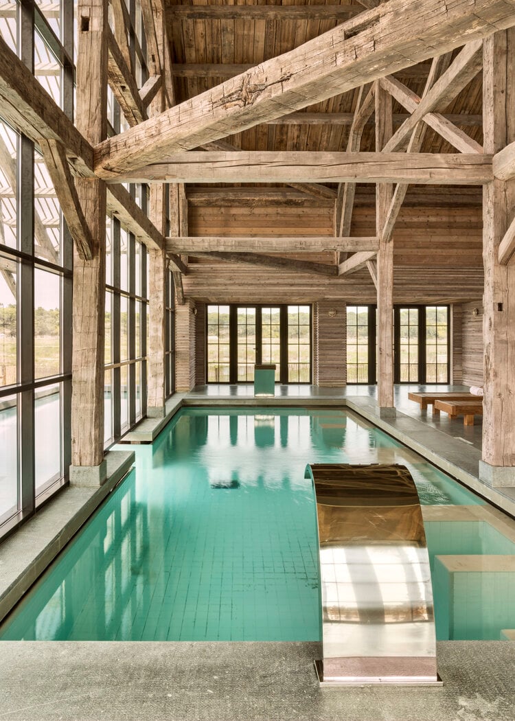 Oryza Spa, Europe — timber-frame indoor pool with turquoise water, exposed beams, tall casement windows, and natural light reflecting on polished concrete deck.