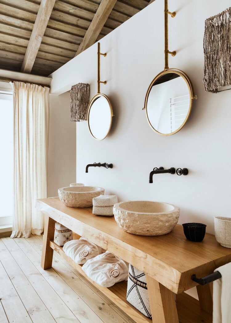 Quinta da Comporta, Europe — Minimalist bathroom with natural stone vessel sinks, brass-framed mirrors, wooden vanity, exposed beam ceiling, and neutral earth-tone palette.