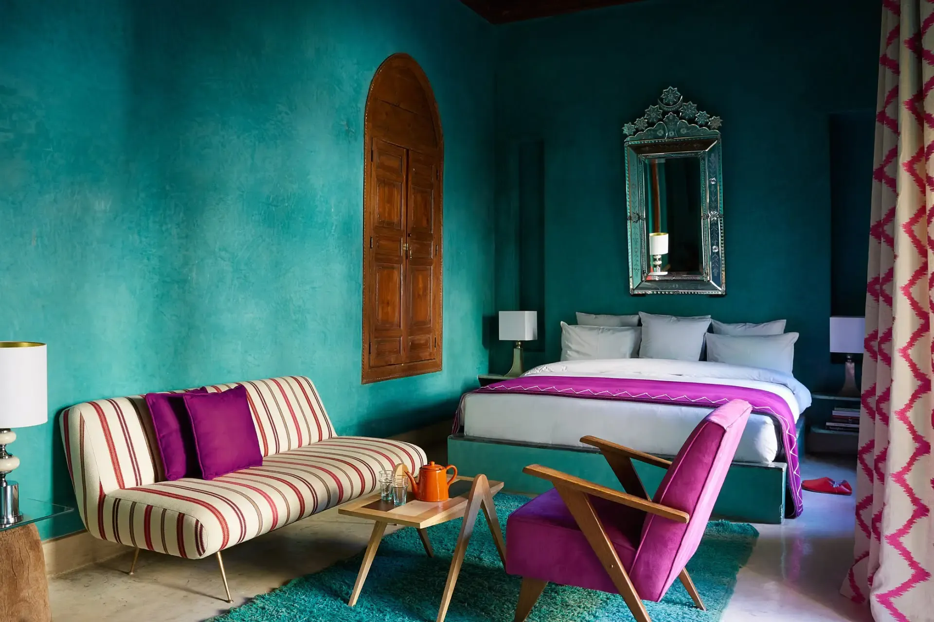El Fenn, Marrakech, Morocco — Turquoise bedroom with striped sofa, magenta accents, arched wooden door, ornate mirror, and mid-century modern furnishings.