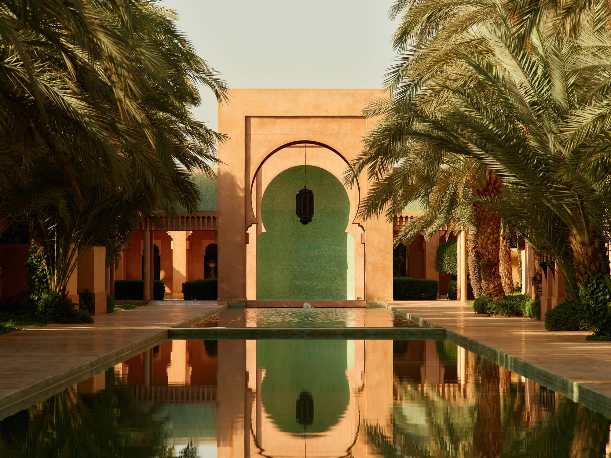 Amanjena, Marrakech, Morocco — Geometric courtyard with reflecting pool, arched green doorway, ochre walls, and palm trees framing Moroccan architecture in golden light.