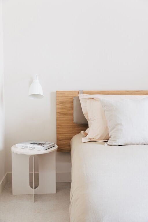 Plácido y Grata, Europe — Minimalist bedroom corner with natural wood headboard, white pendant lamp, cream linens, and cylindrical nightstand in serene neutral palette.