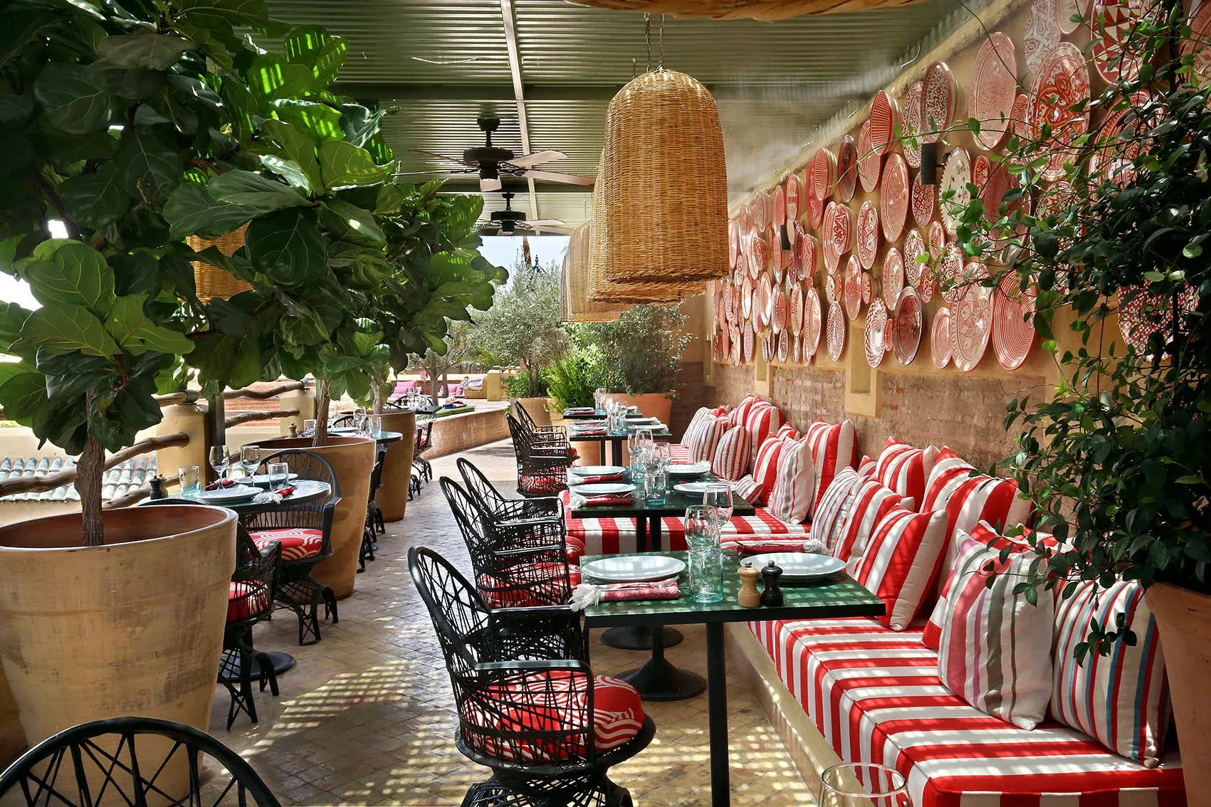 El Fenn Restaurant, Africa — Red-and-white striped banquette seating with black chairs beneath woven pendant light, terracotta planters, decorative plates, and lush greenery in covered courtyard.