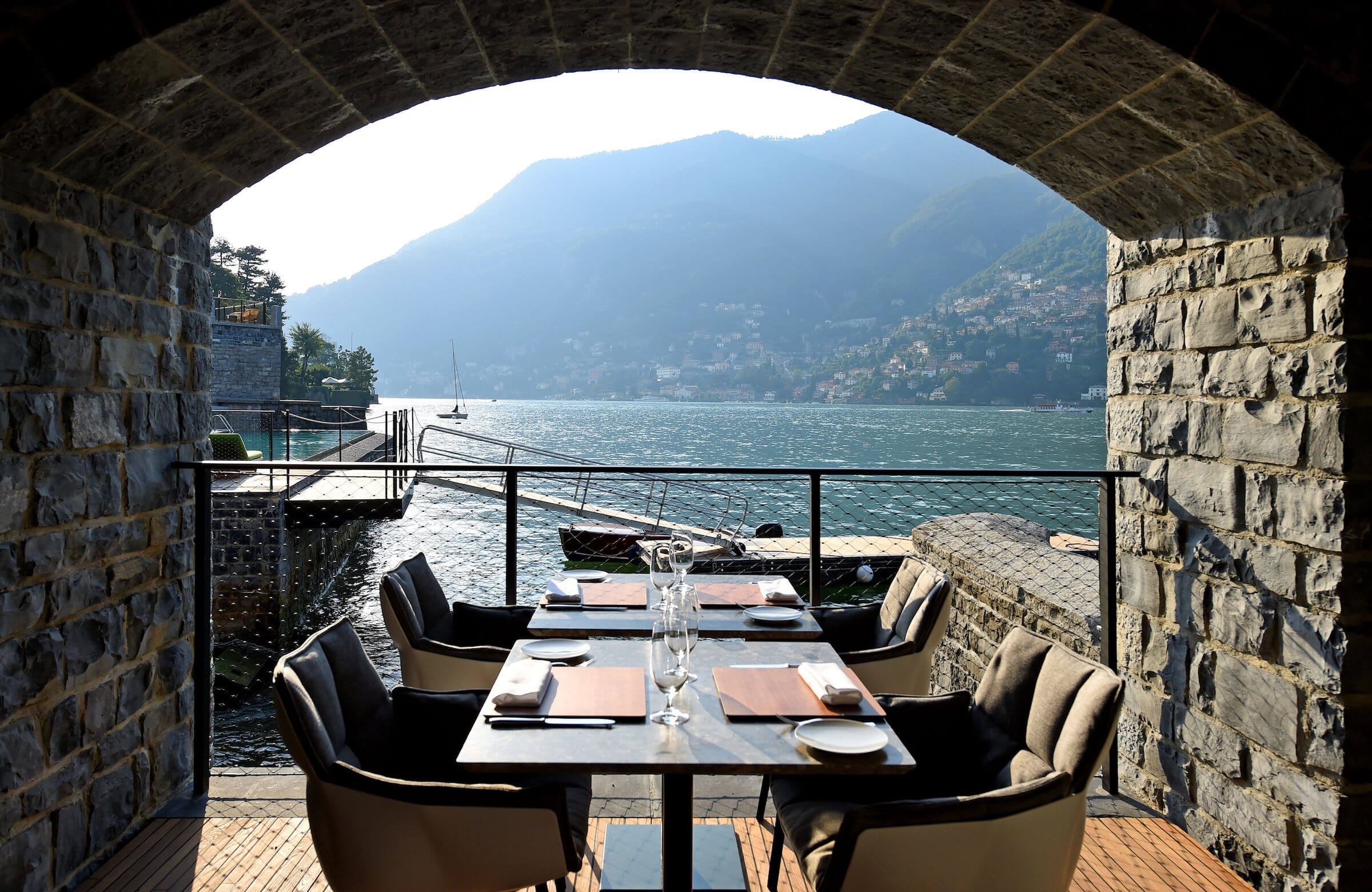 il Sereno Lago di Como, Lake Como, Italy — Stone archway opening onto lakeside dining terrace with mountain views, modern furniture, sailboat on water.