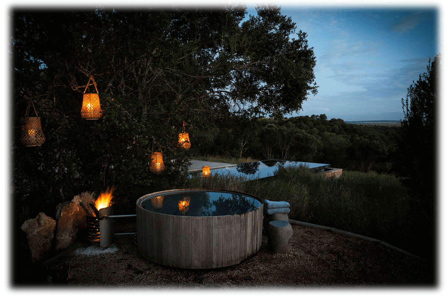 Arijiju Retreat, Africa — Wooden hot tub at dusk with hanging lanterns, fire pit, and waterhole views through acacia trees.