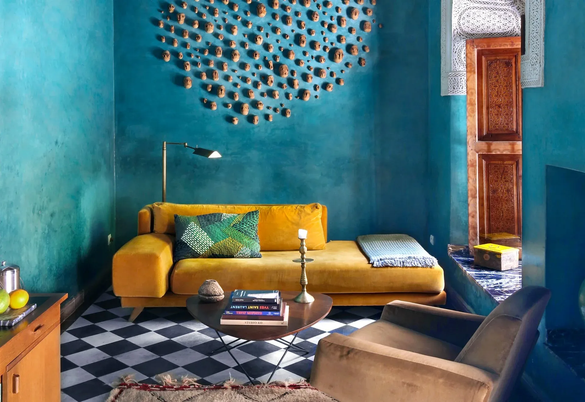 El Fenn, Marrakech, Morocco — turquoise living room with golden velvet sofa, geometric floor, terracotta door, brass floor lamp, and sculptural clay wall installation.