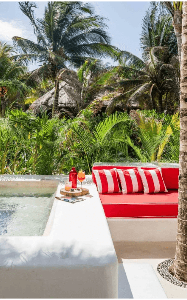Hotel Esencia, Mexico — Red-and-white striped cushions on a waterfront lounger overlooking turquoise lagoon, thatched-roof bungalows, and palm trees.