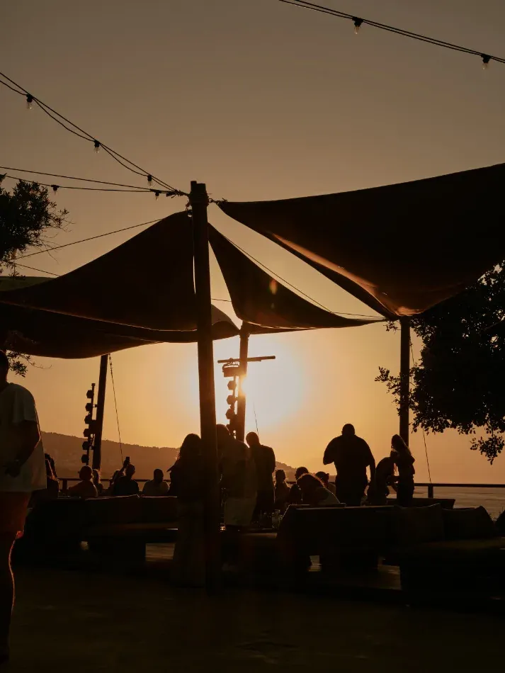 The Terrace, Mediterranean — Candlelit outdoor dining pavilion at golden hour with silhouetted guests, fabric canopy, and coastal water views.