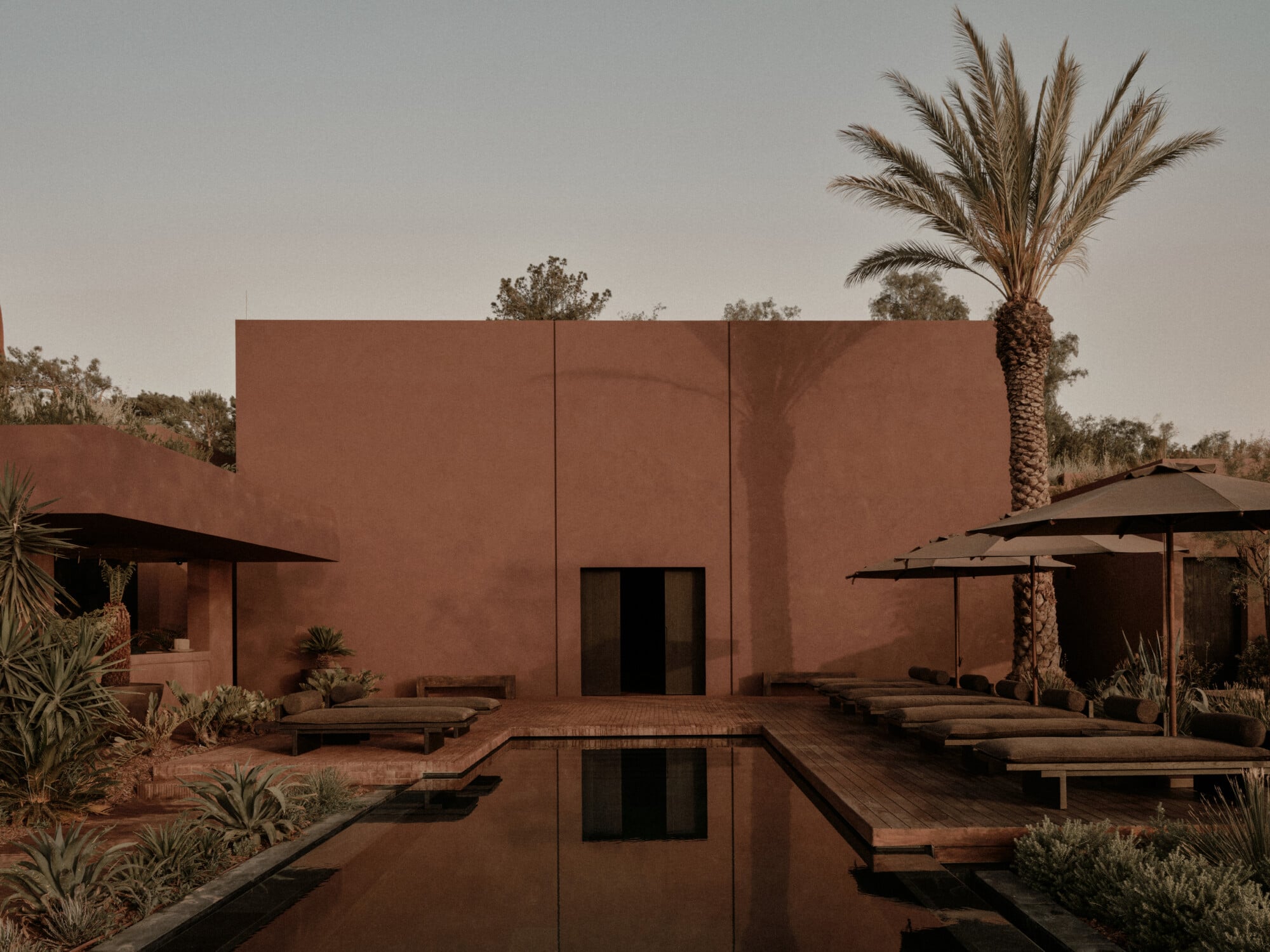 Scorpios Bodrum, Mediterranean — Terracotta courtyard with reflecting pool, minimalist adobe architecture, palm trees, wooden loungers and umbrellas at sunset.