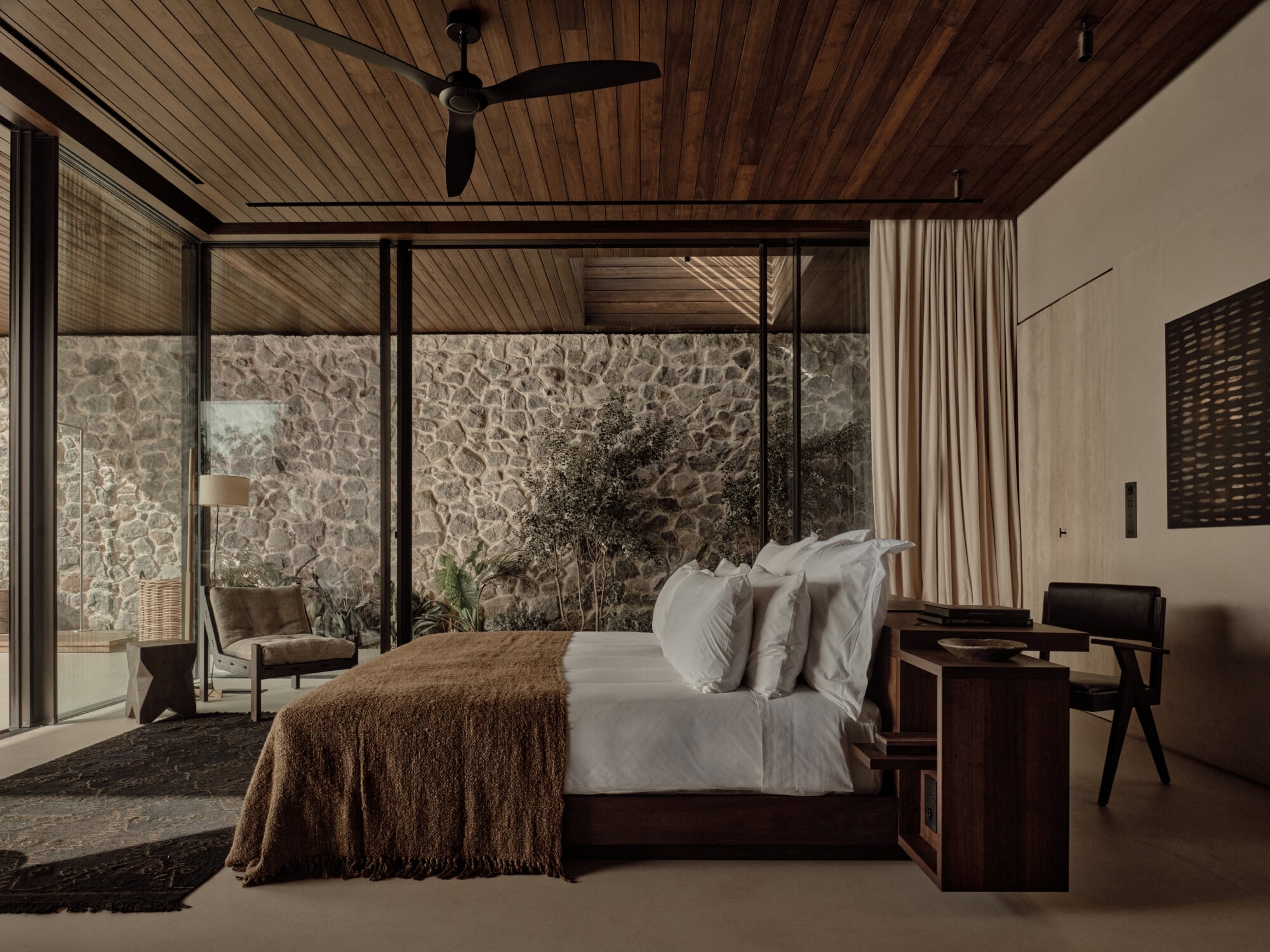 Scorpios Bodrum, Mediterranean — Modern bedroom with wooden ceiling, stone feature wall, floor-to-ceiling glazing, dark wood furniture, and minimalist neutral palette overlooking landscape.