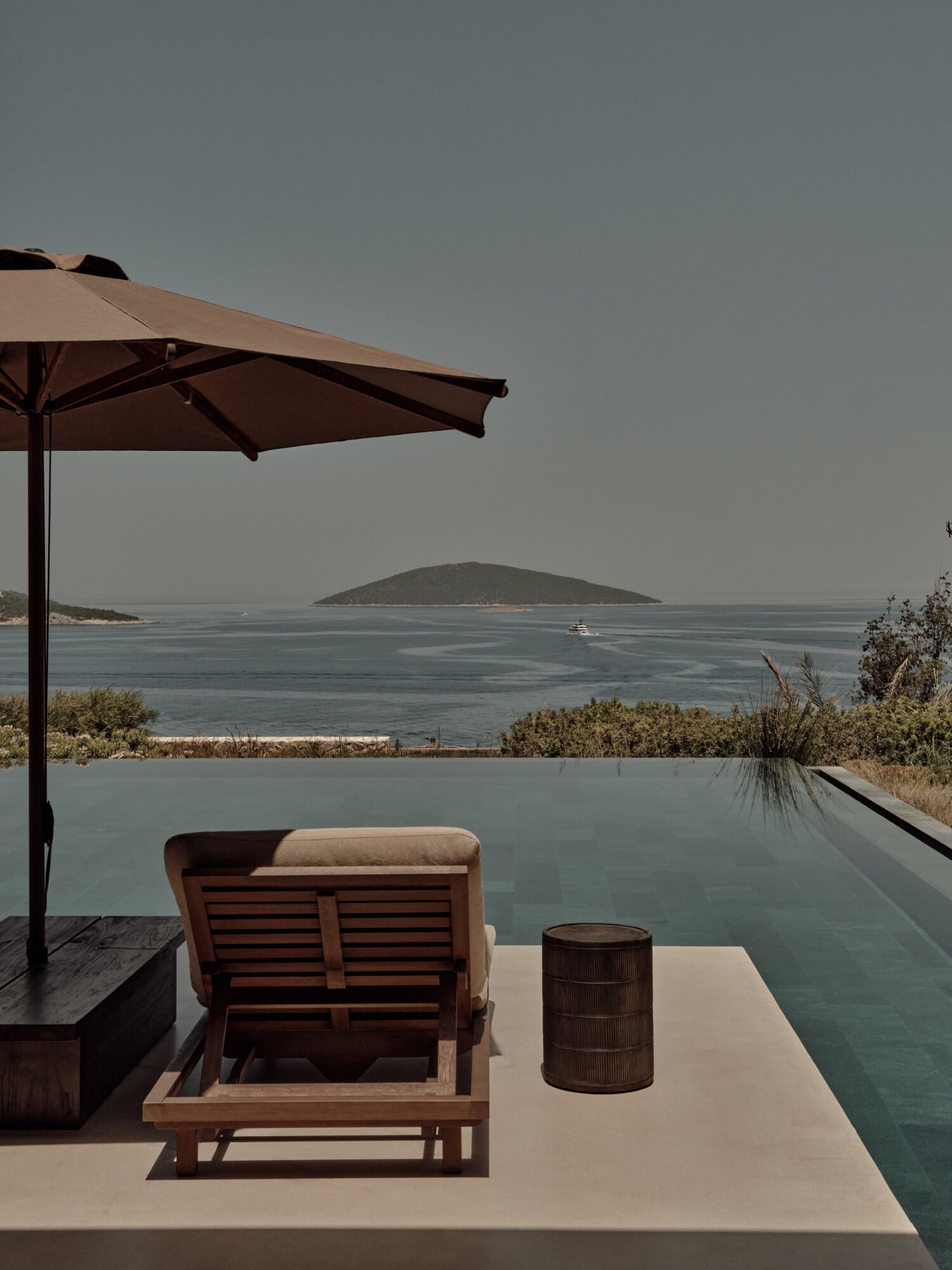 Scorpios Bodrum, Mediterranean — Minimalist poolside terrace with wooden lounge chair, bronze umbrella, and infinity pool overlooking Aegean Sea island vista.
