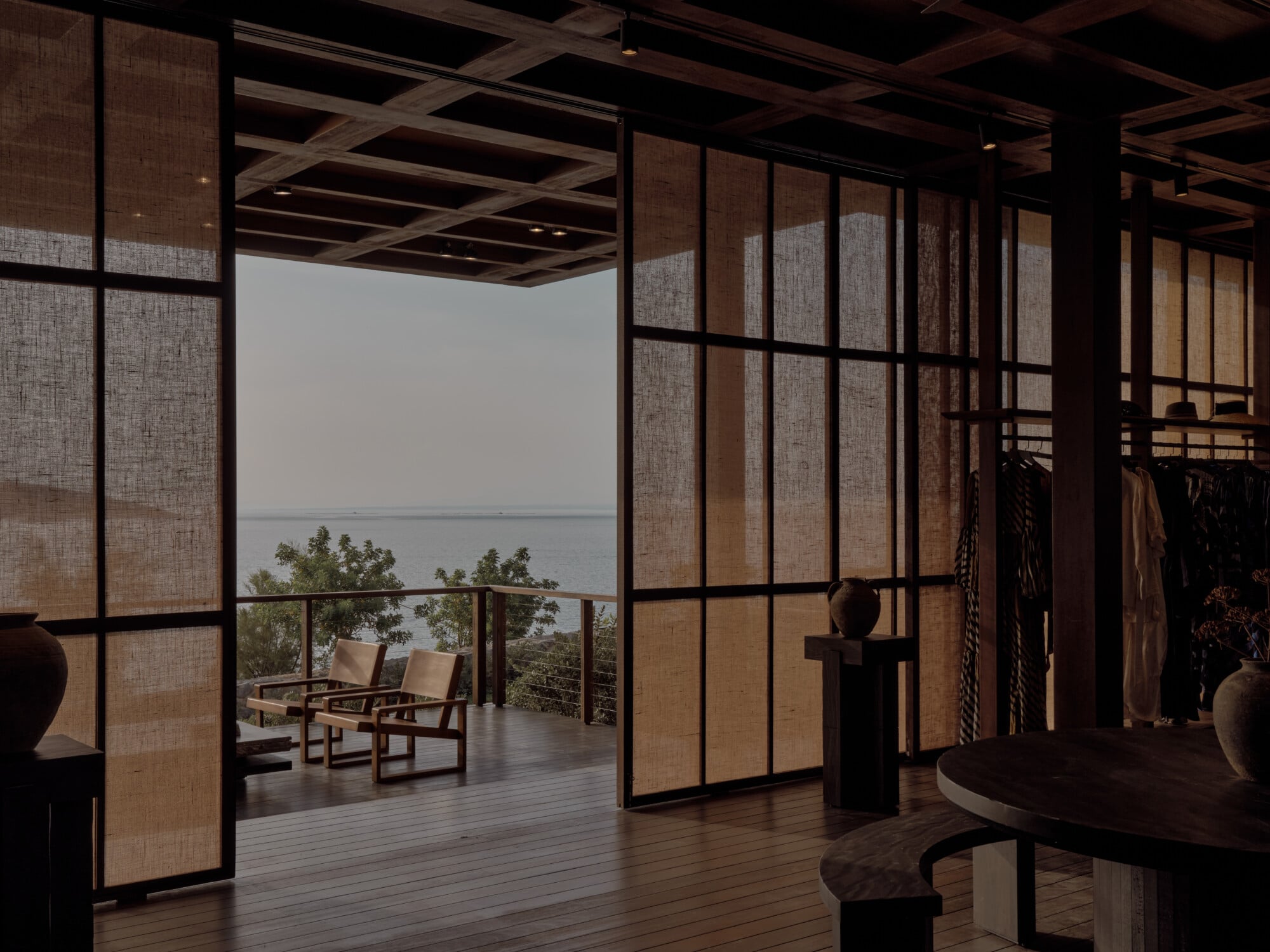 The Scorpios Bazaar, Mediterranean — Linen room dividers frame coastal views, wooden ceiling beams and dark flooring create warm minimalist retail space with terraced seating area overlooking water.