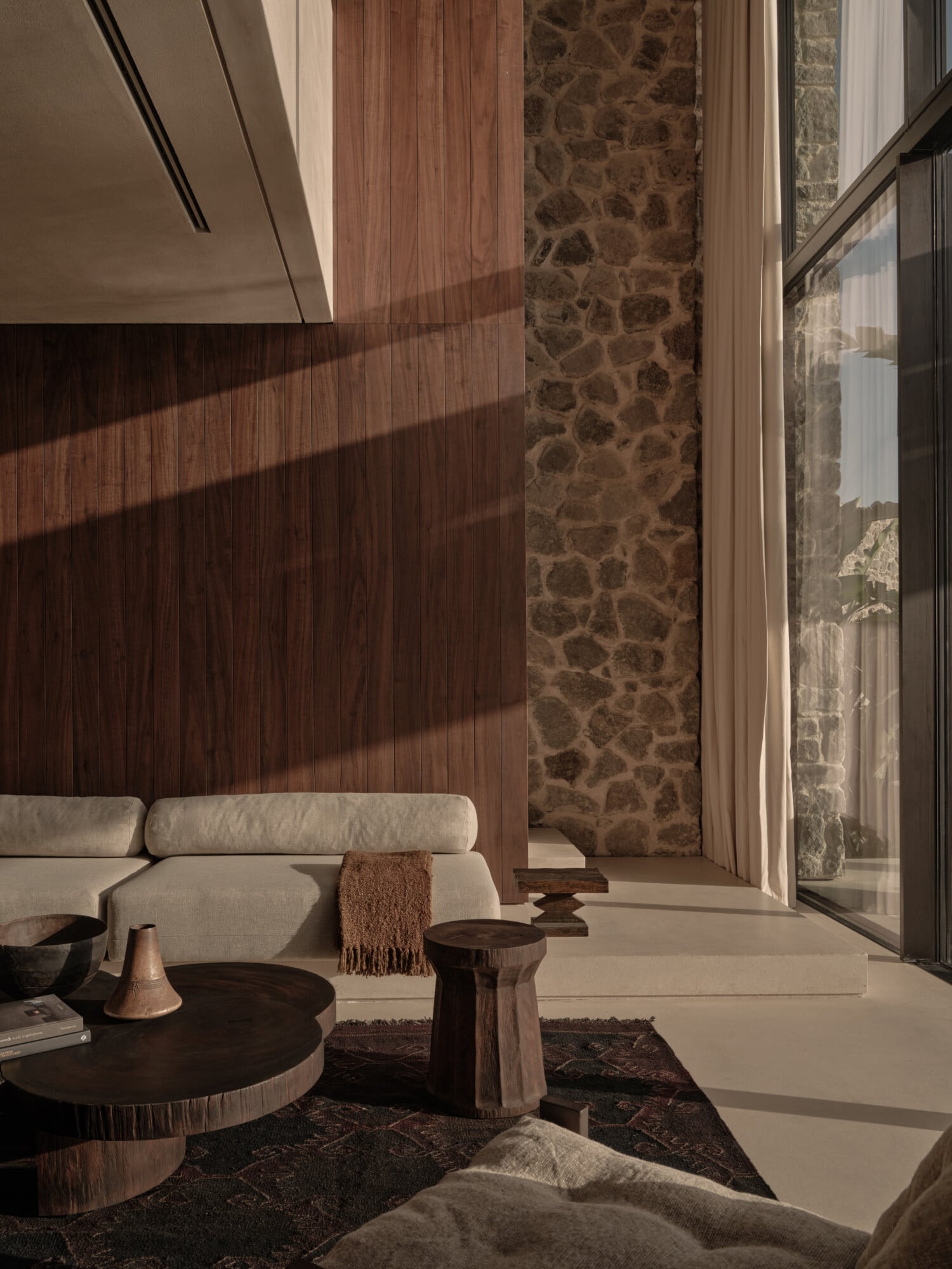 Scorpios Bodrum, Mediterranean — Minimalist bedroom with warm wood accent wall, stone pillar, cream curved sofa, natural light, modern luxury design.