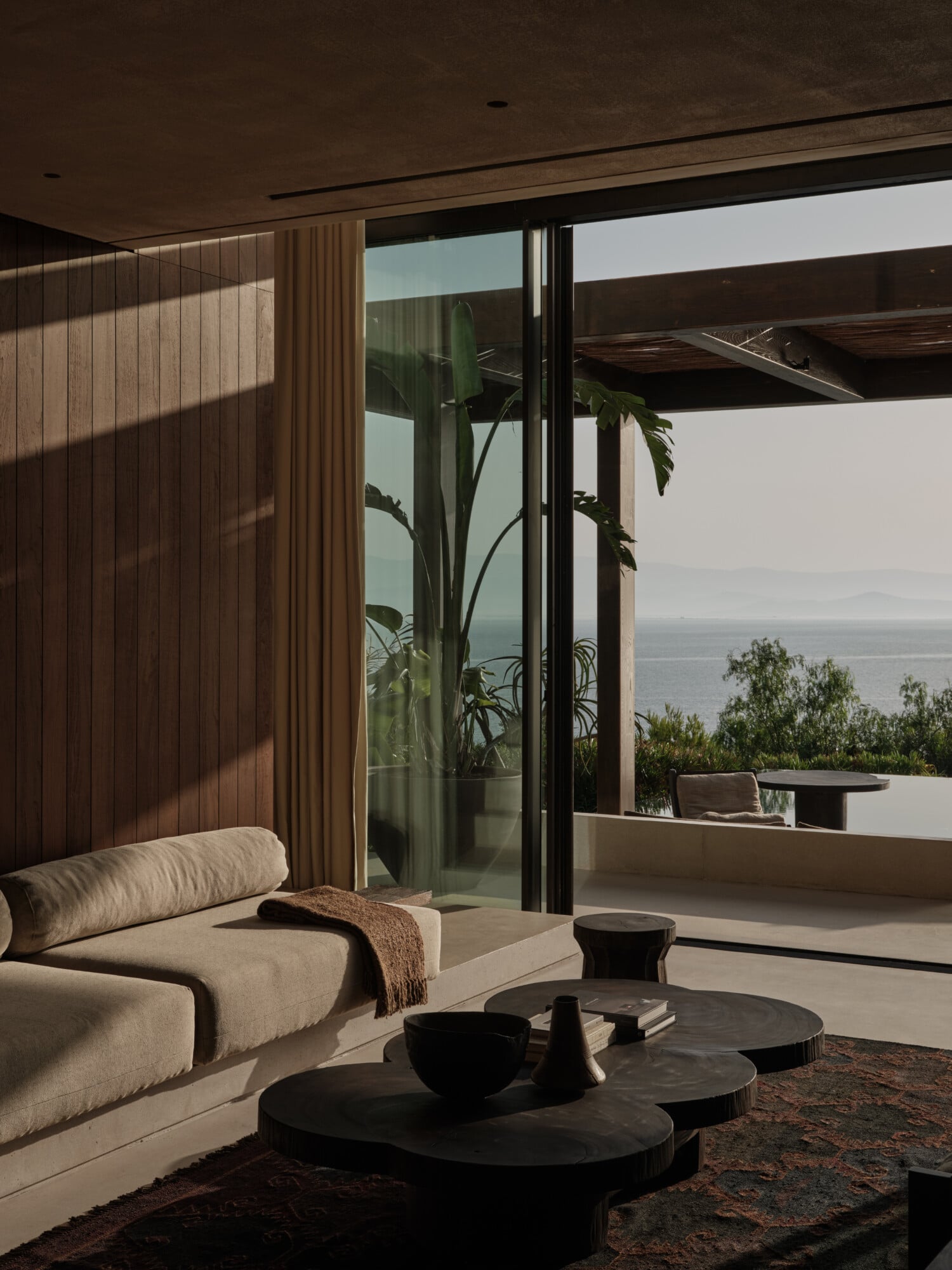 Scorpios Bodrum, Mediterranean — Minimalist villa interior with cream curved sofa, black stone tables, floor-to-ceiling windows, vertical wood paneling, sea view terrace with plants and morning light.