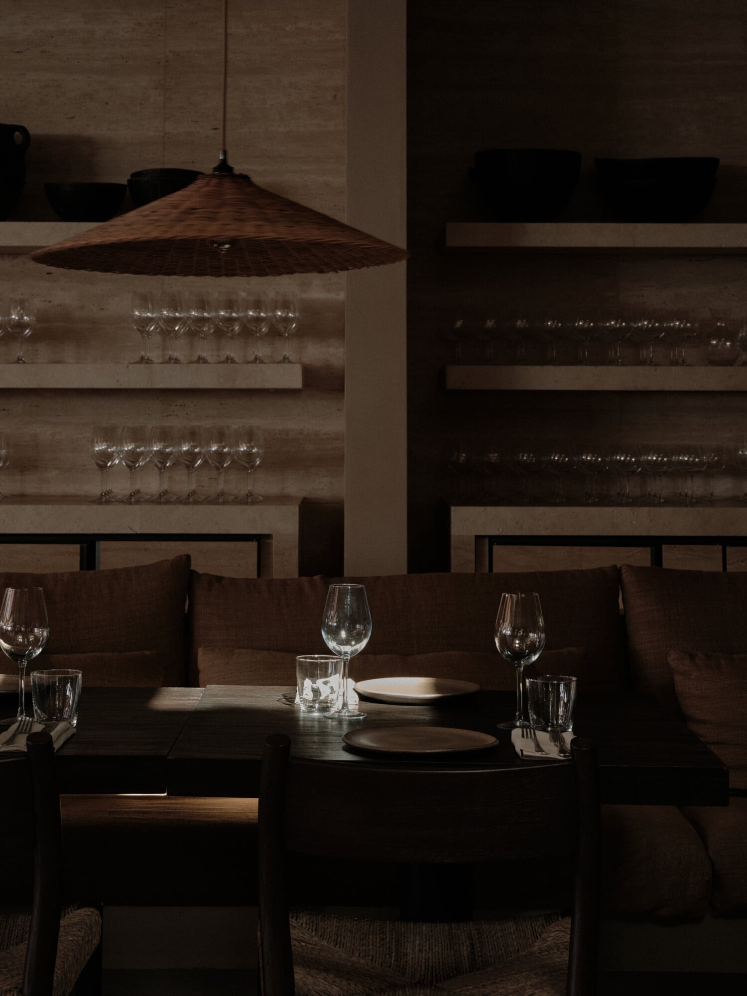 Scorpios Bodrum, Mediterranean — Minimalist dining room with woven pendant lamp, concrete walls, dark wood tables, wine glasses, and soft natural light casting geometric shadows.