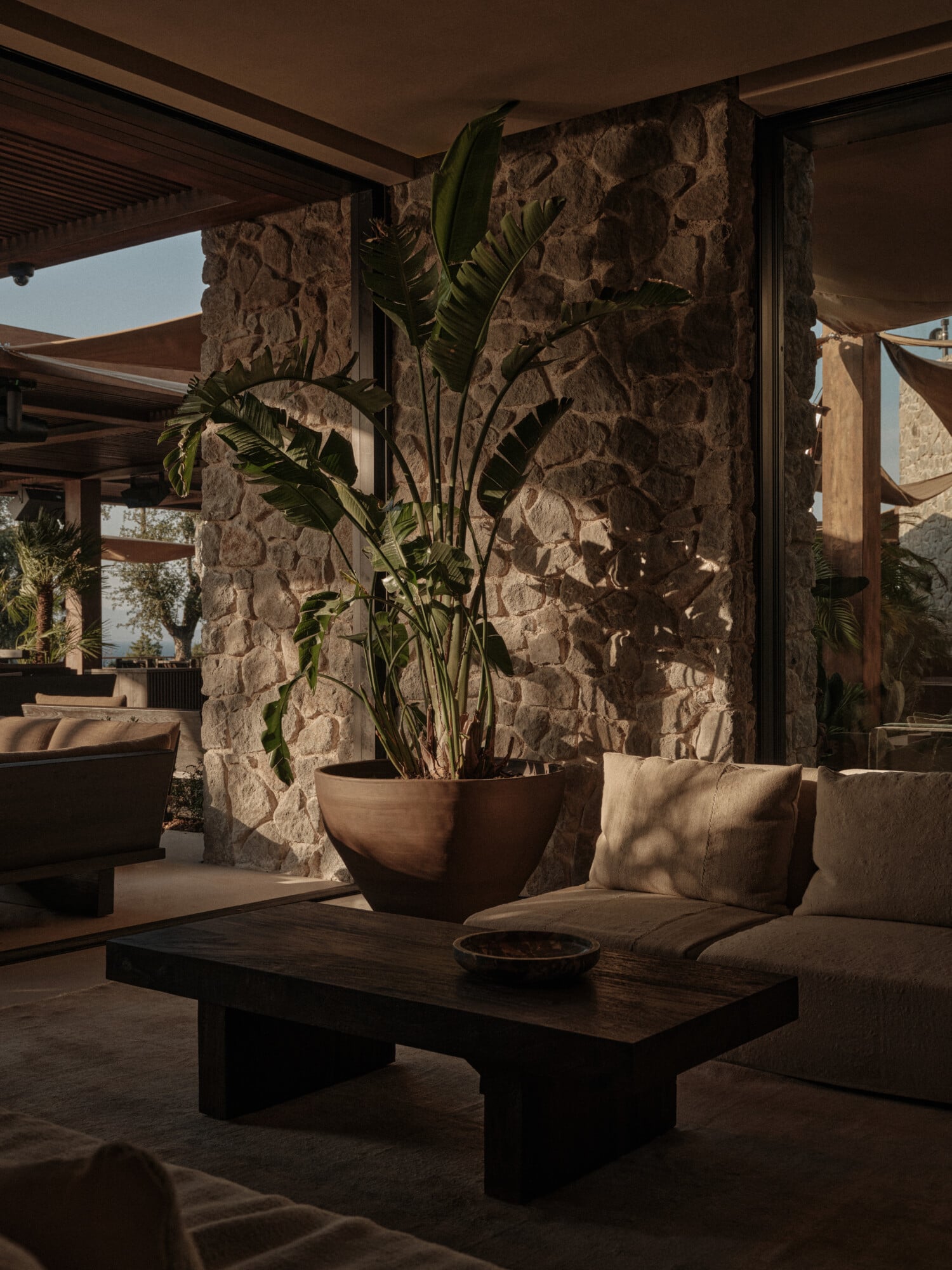 Scorpios Bodrum, Mediterranean — Stone terrace with dark wooden bench, linen pillows, potted palm plant, and textured masonry wall under natural sunlight.