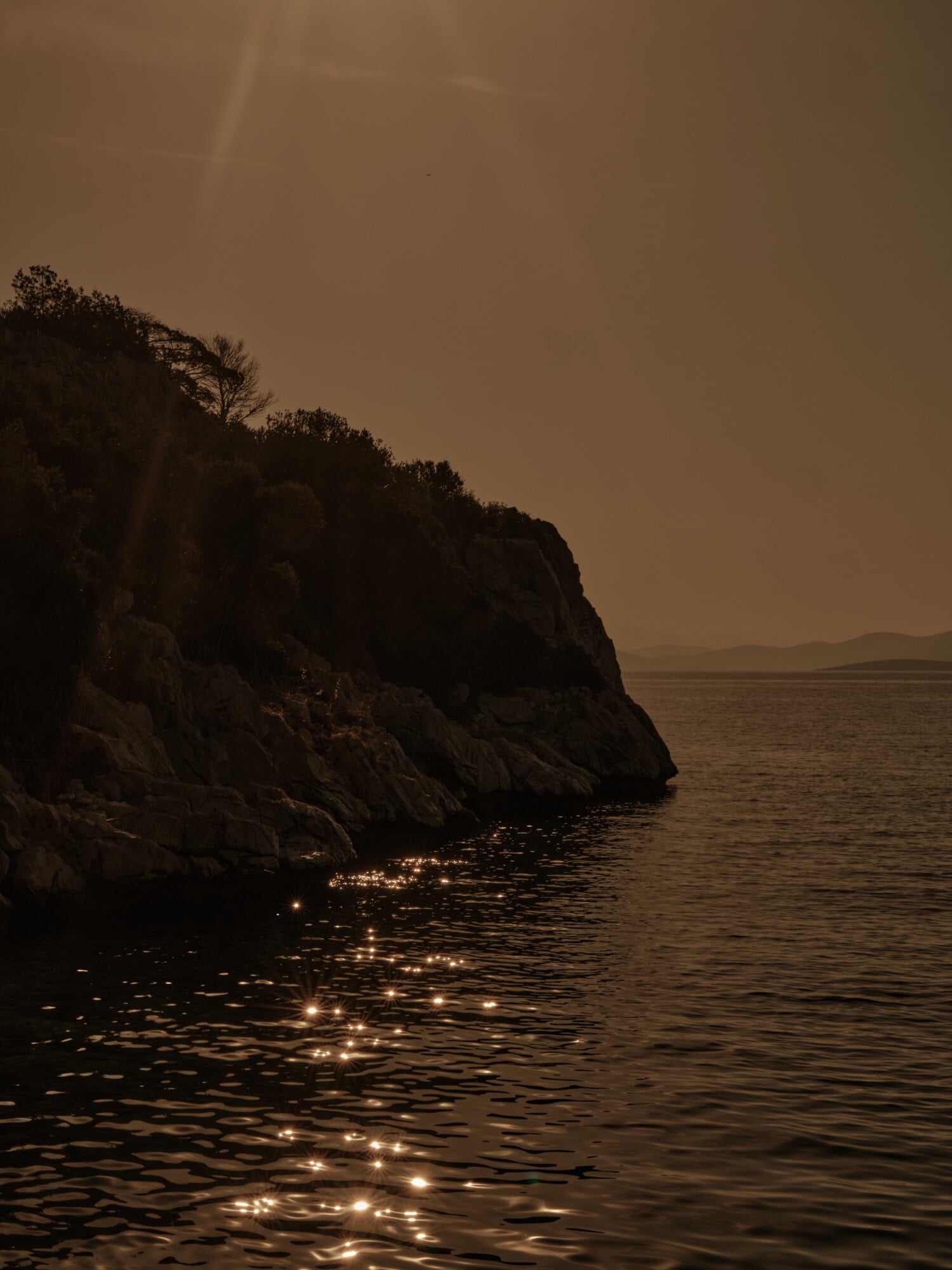 Scorpios Bodrum, Mediterranean — Rocky clifftop at dusk with golden light reflecting across calm water, silhouetted vegetation, hazy horizon.