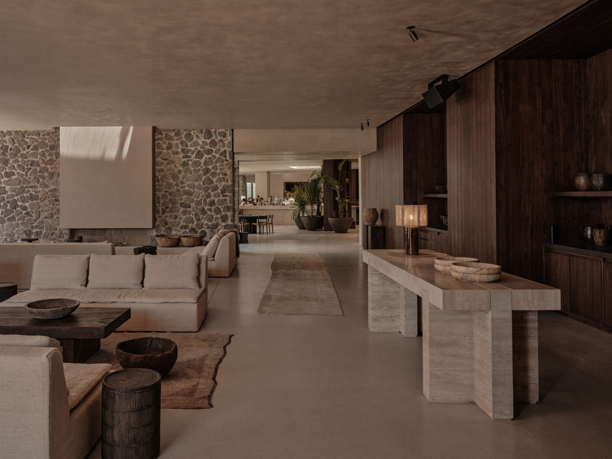 Scorpios Bodrum, Mediterranean — Minimalist lounge with cream modular sofa, travertine console, dark wood cabinetry, stone accent wall, and natural light through skylights.