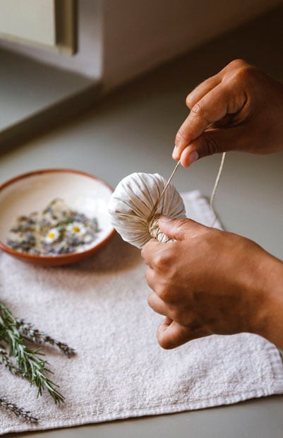 Susanne Kaufmann Spa Barrocal, Europe — hands tying herbal tea sachet over ceramic bowl with dried botanicals on linen, natural light wellness ritual.