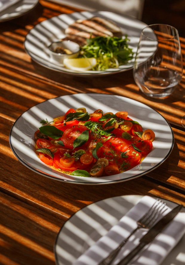 Pool Bar, Europe — Sunlit wooden table set with tomato and basil salad, striped shadows across white plates, minimalist fine dining presentation.