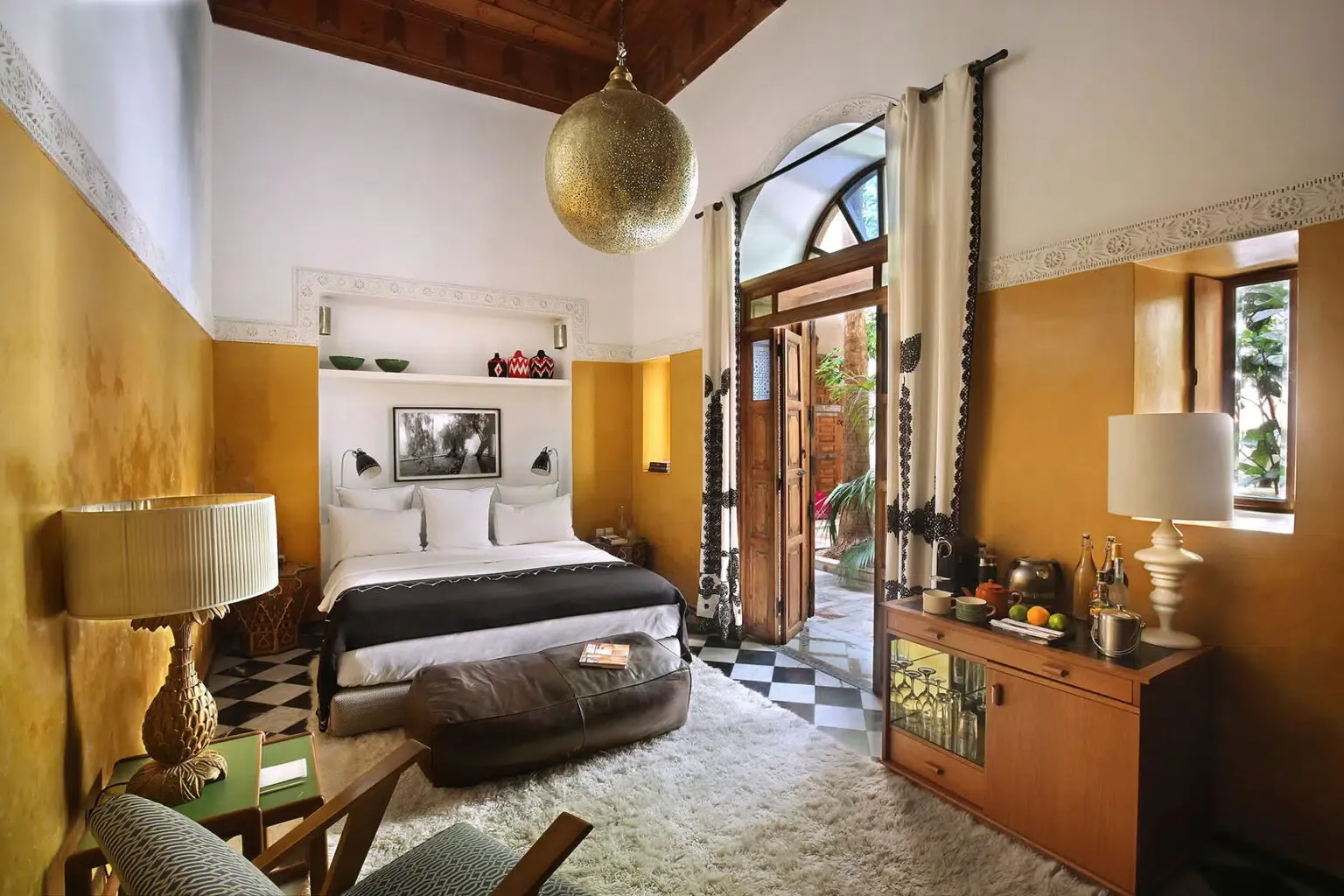 El Fenn, Marrakech, Morocco — Mustard-yellow bedroom with arched doorway, brass pendant light, black-and-white floor tiles, wooden beamed ceiling, and view to courtyard.
