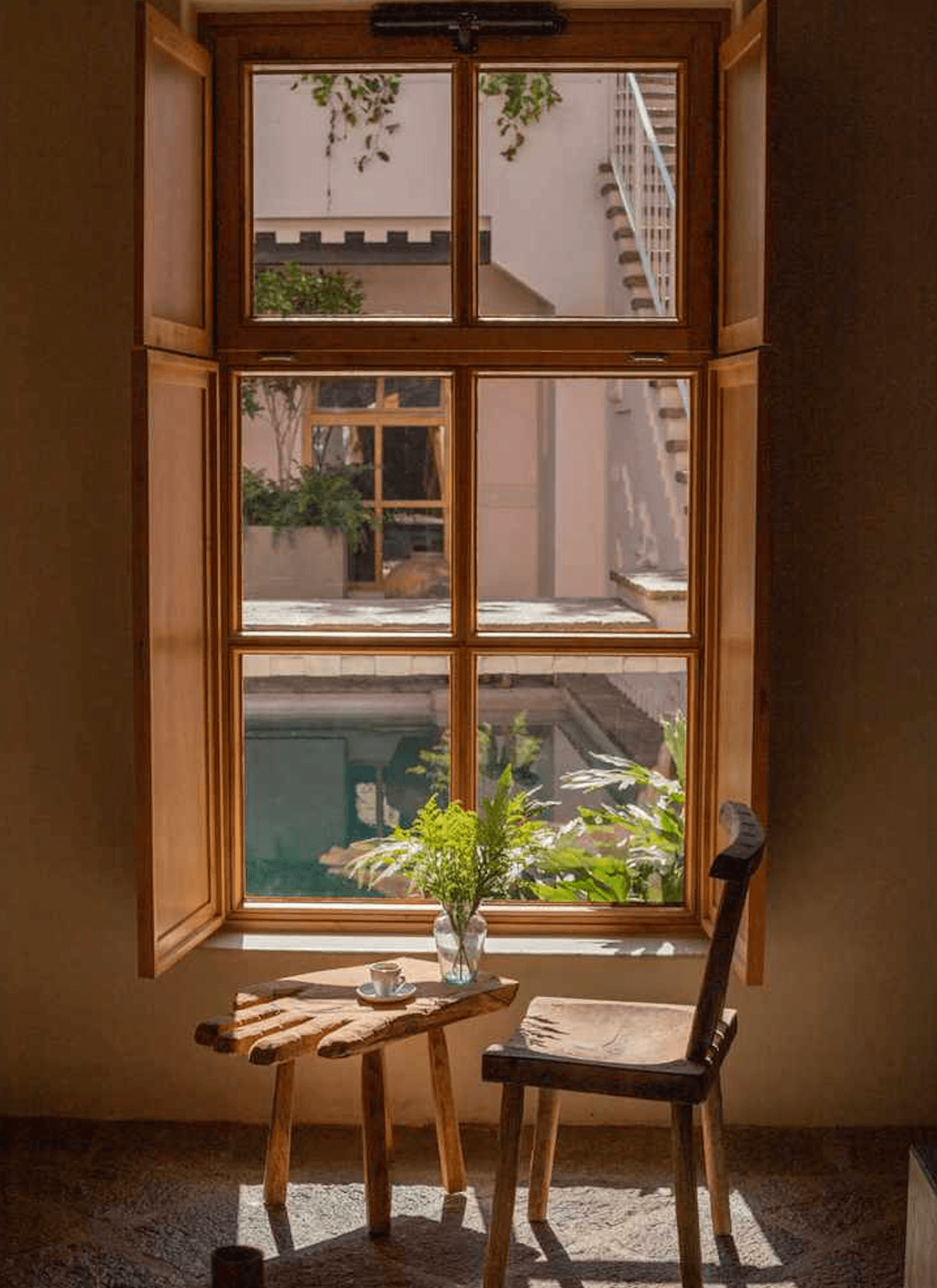 La Valise San Miguel, Mexico — Wooden-framed window opening onto courtyard, natural light illuminating simple chair, small table with fresh flowers, and coffee cup in minimalist interior.