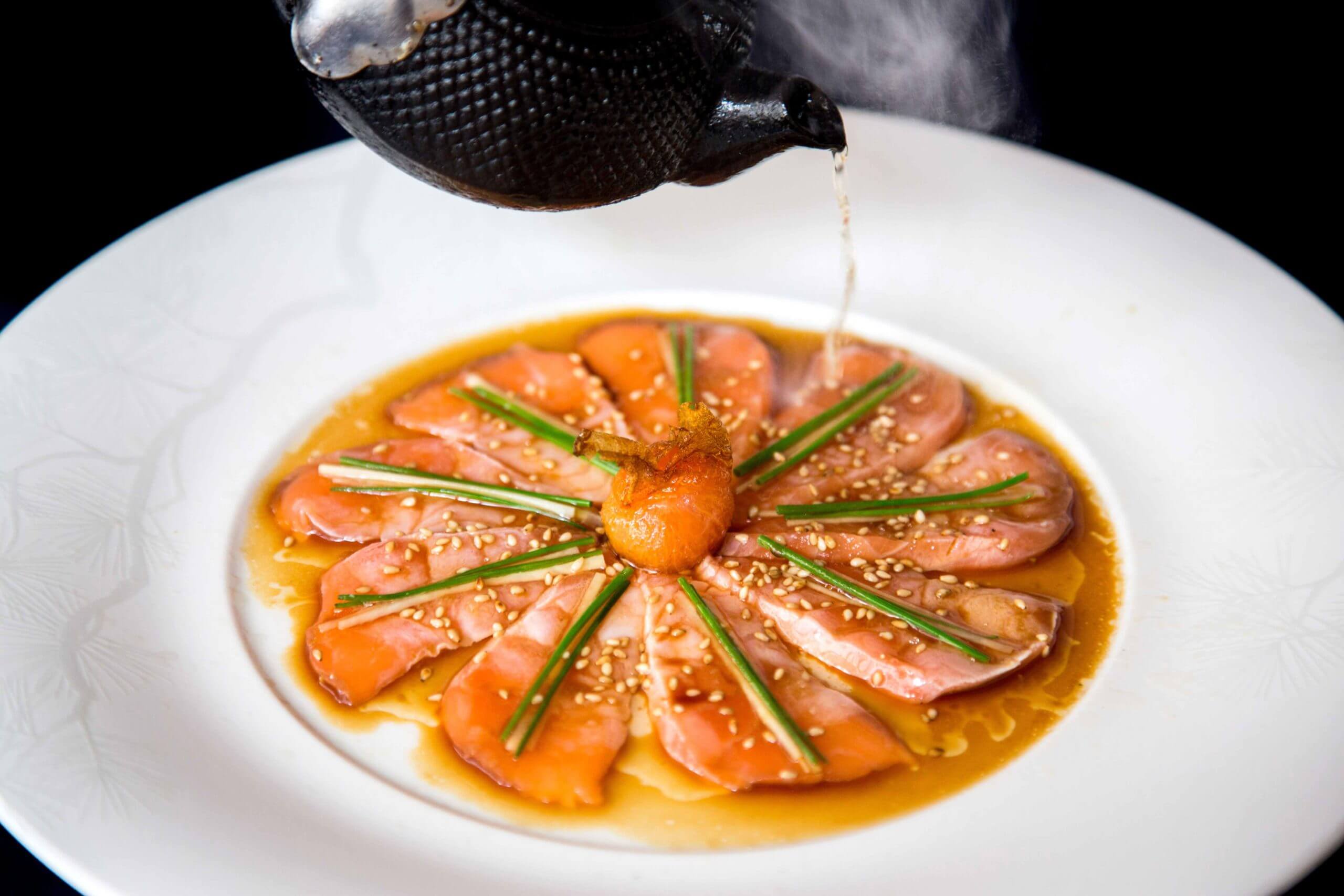 Nobu Warsaw, Europe — Seared salmon sashimi arranged in radial pattern with sesame seeds, chives, and amber dashi broth poured tableside on white plate.