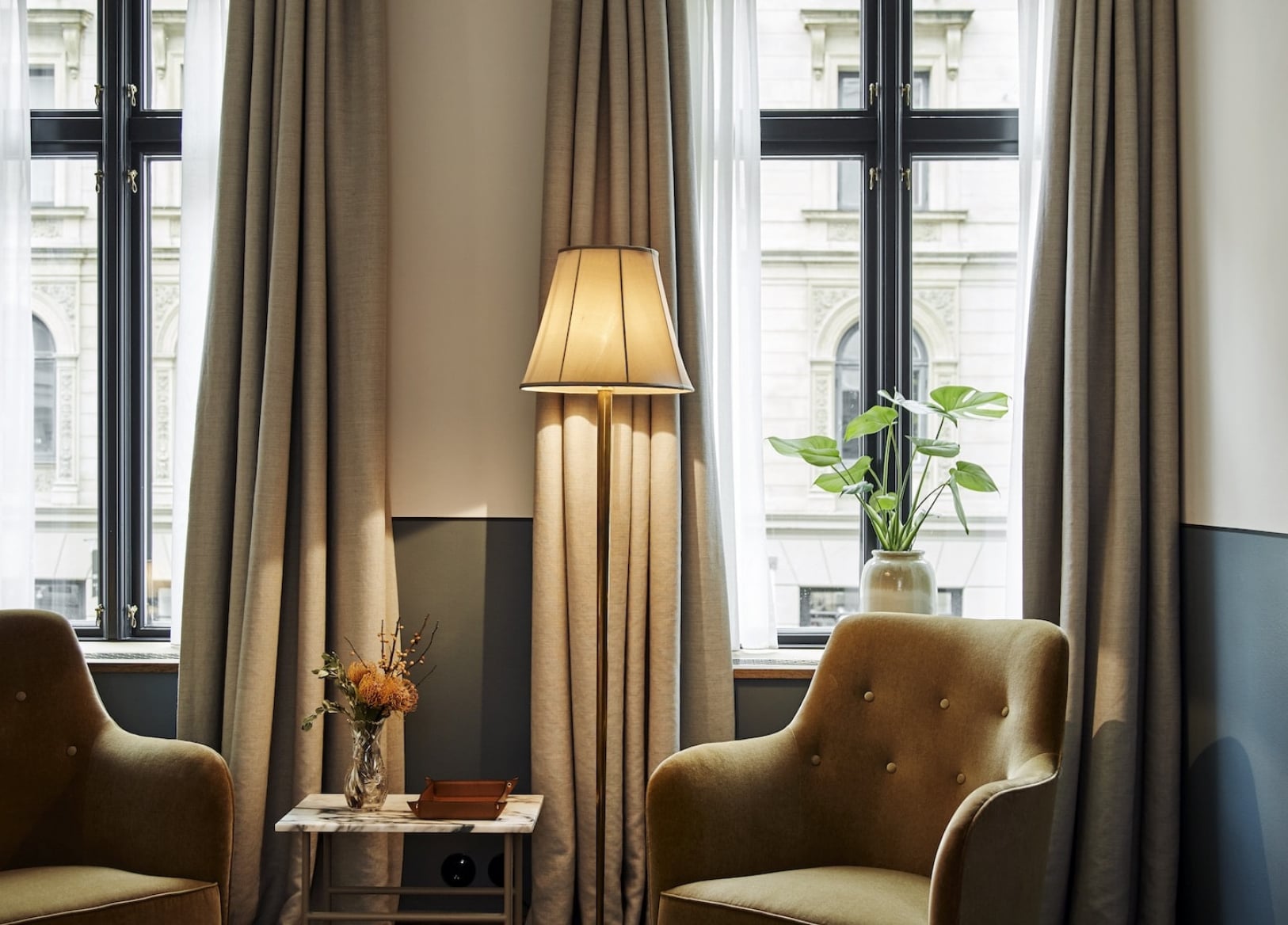 Hotel Sanders, Europe — Elegantly appointed seating area with taupe upholstered chairs, brass floor lamp, marble side table, floor-to-ceiling windows with natural drapes overlooking classical architecture.