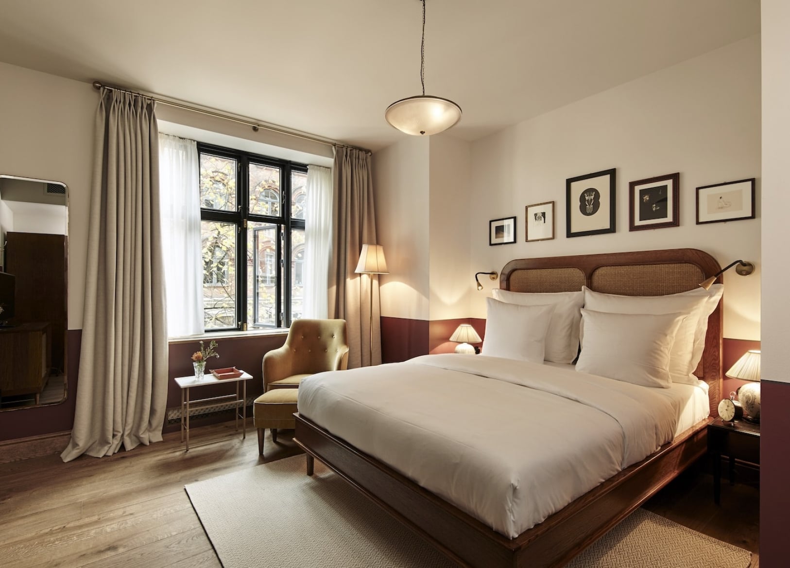 Hotel Sanders, Europe — Elegant bedroom with dark wood bed, cream linens, burgundy accent wall, pendant lighting, and black-framed gallery wall above headboard.