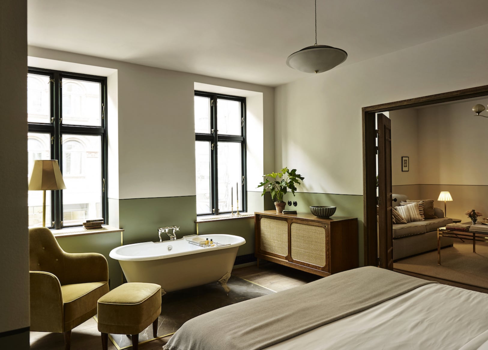 Hotel Sanders, Europe — Luxe bedroom-bathroom suite with sage-green accent wall, freestanding soaking tub, mid-century furniture, and natural light from black-framed windows.