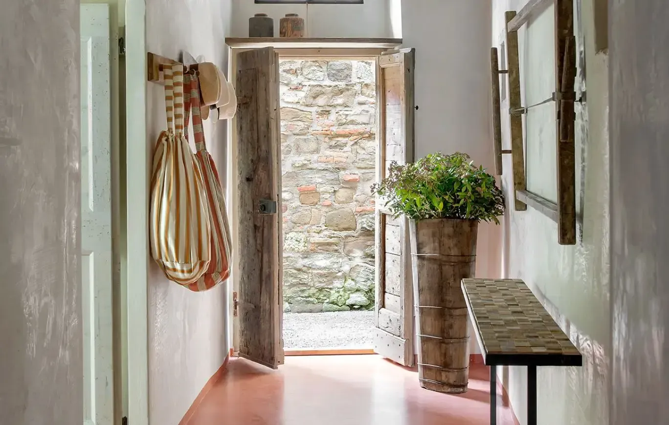 Monteverdi, Europe — Rustic hallway with whitewashed walls, wooden beamed door, terracotta floor, potted greenery, and open courtyard beyond stone facade.
